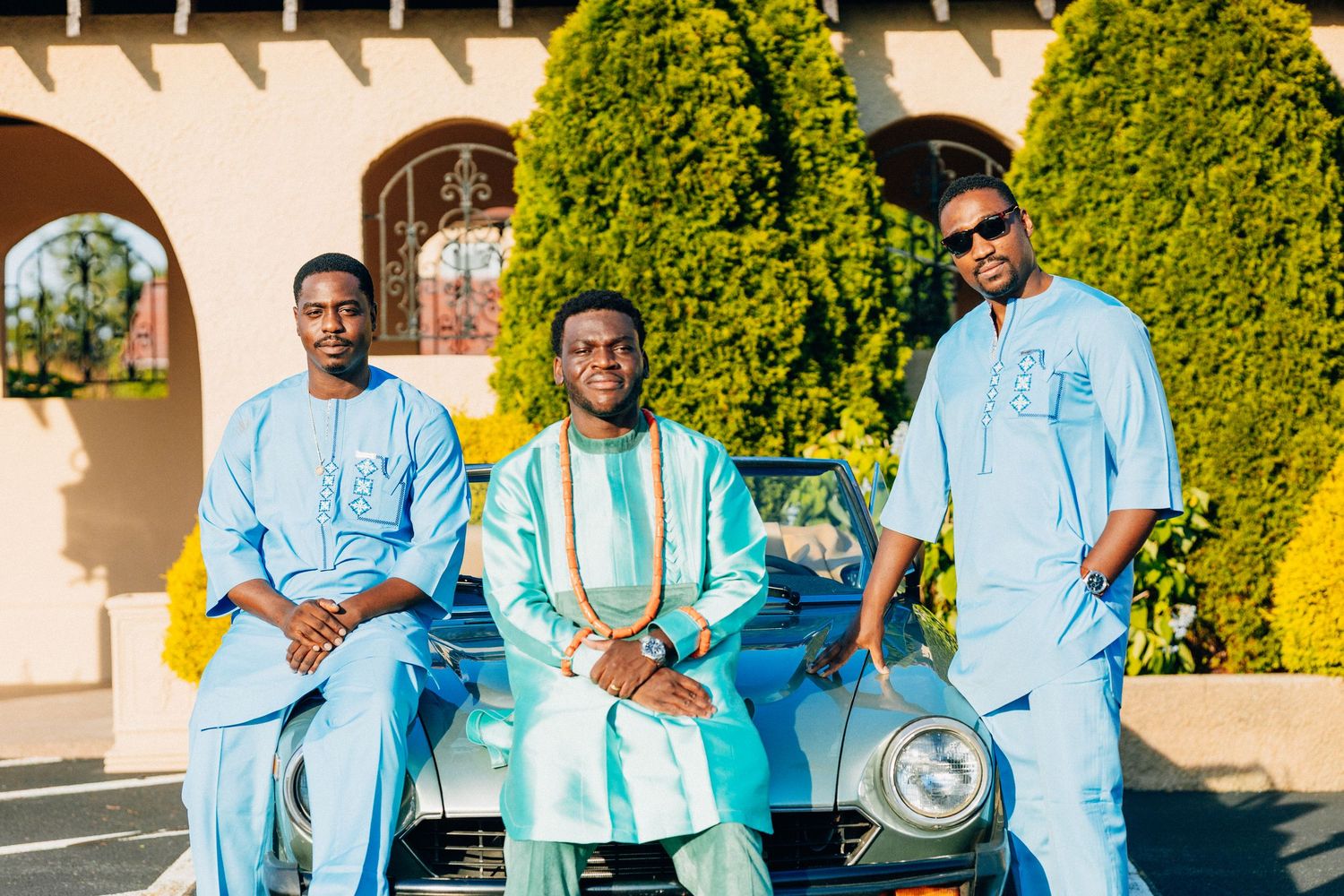 Three men in African traditional attire pose with vintage blue car outdoors.