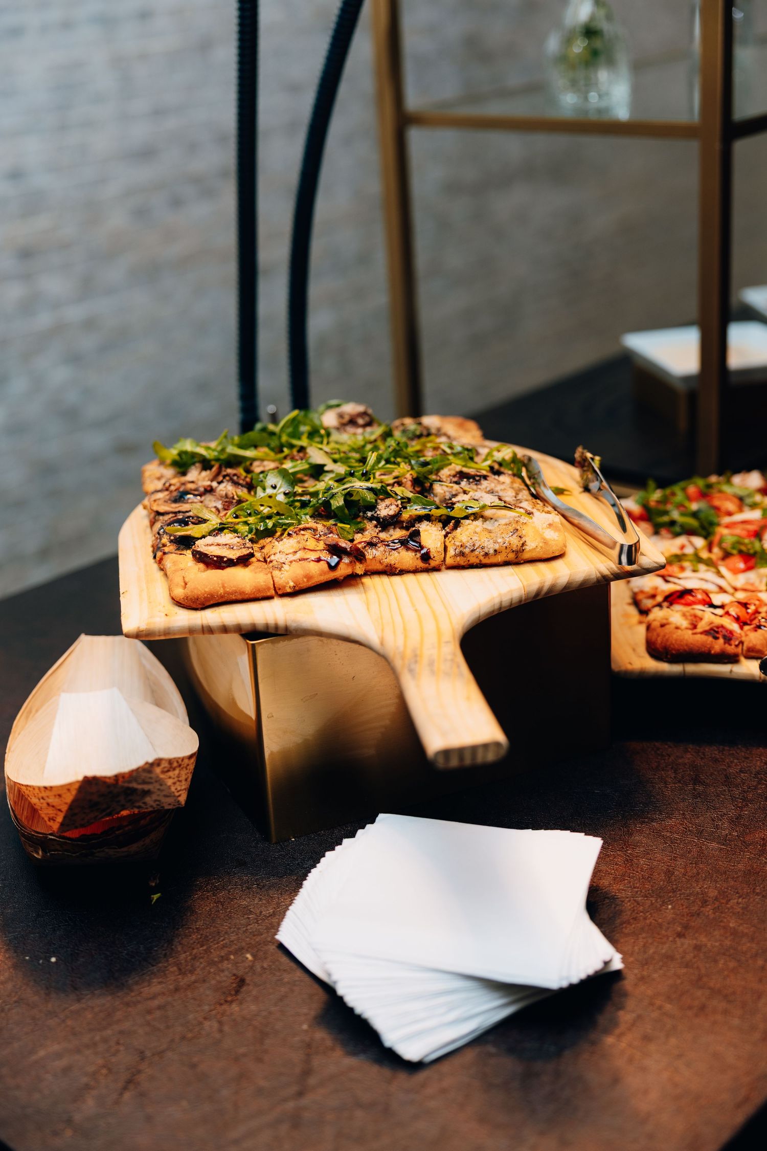 Two flatbread pizzas topped with arugula and vegetables displayed on wooden boards at a catering station.
