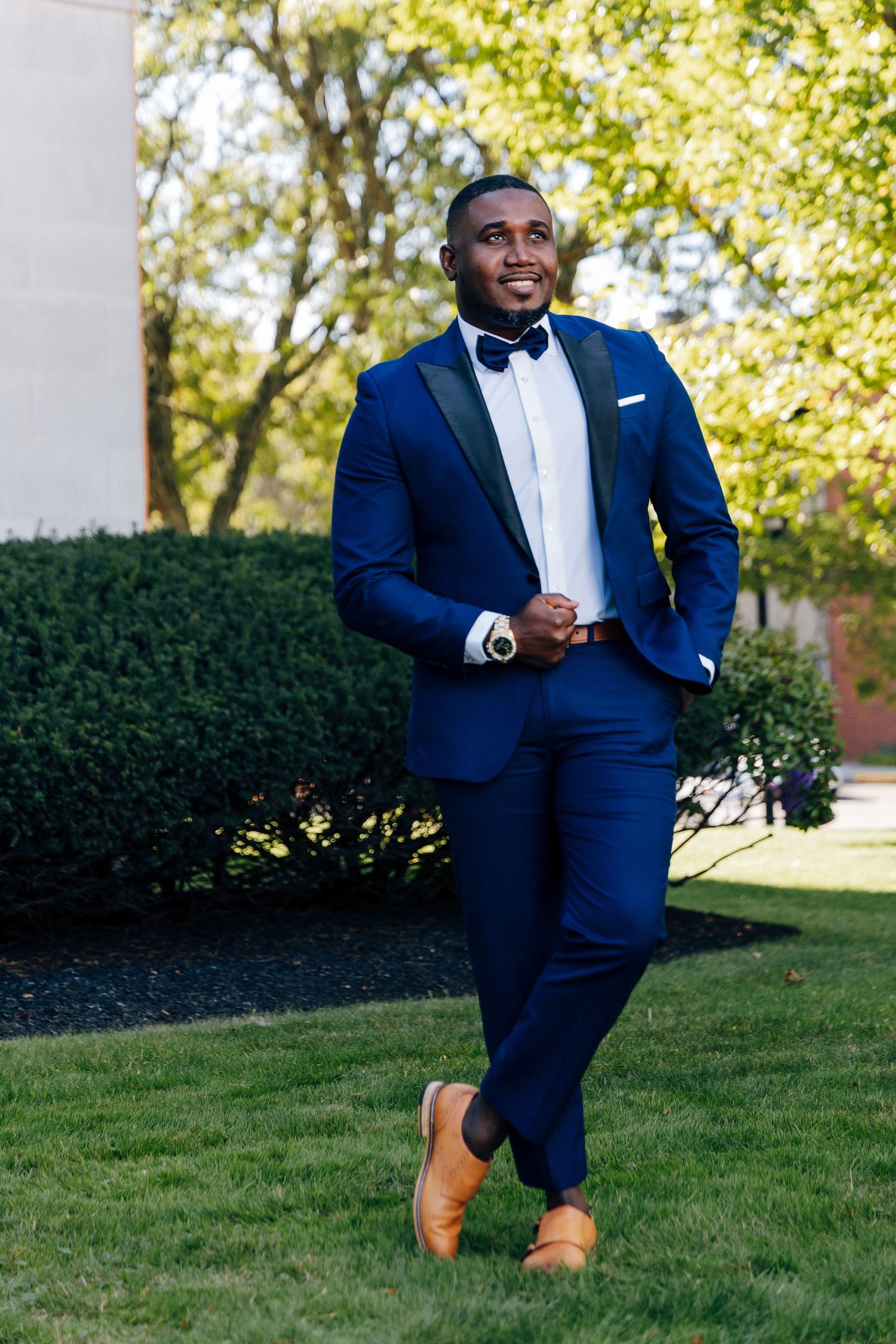 Dapper groom in navy blue tuxedo with bow tie and tan oxford shoes posing on green lawn.