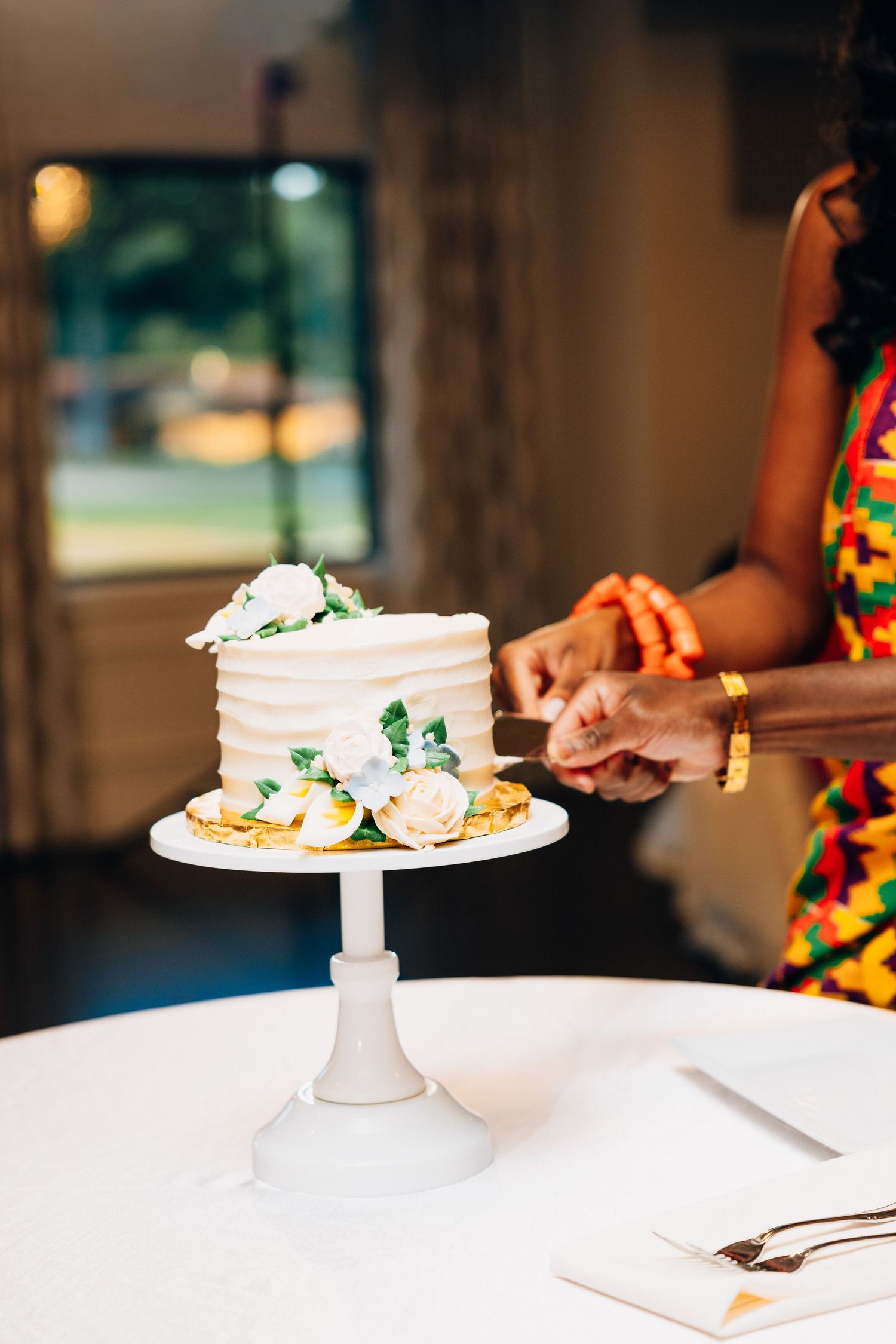 Bride cutting a white single-tier wedding cake decorated with flowers on a pedestal.