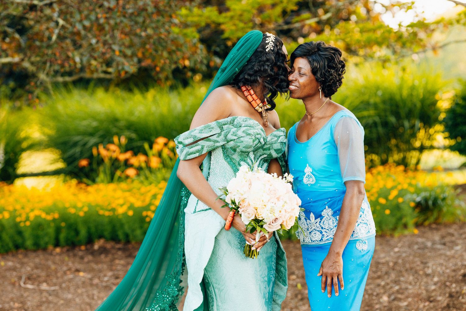 Two women in elegant teal and blue traditional gowns share a tender moment outdoors surrounded by lush greenery.
