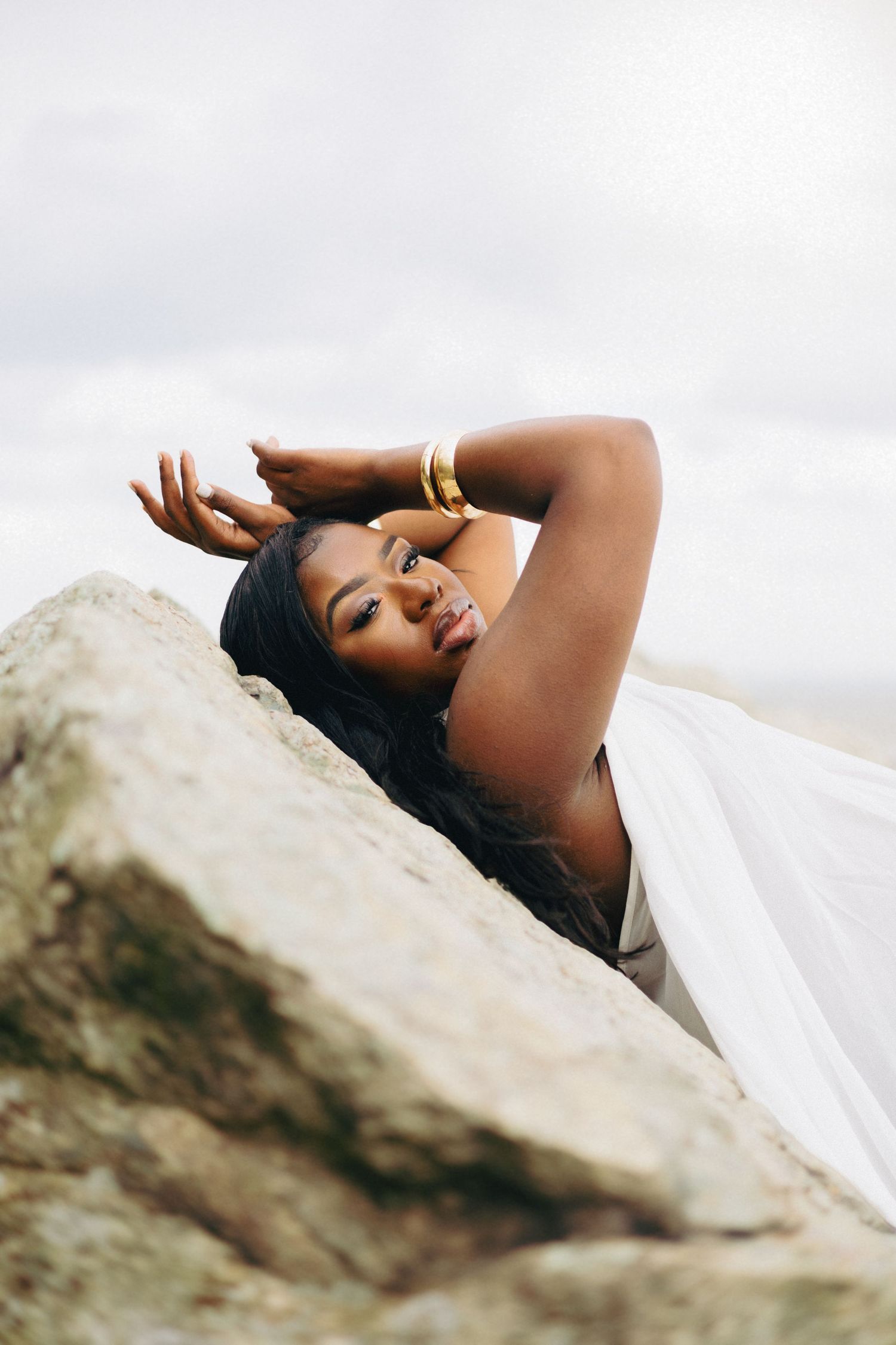 A Black woman in a white dress and gold bracelets poses gracefully on rocks against a cloudy sky.