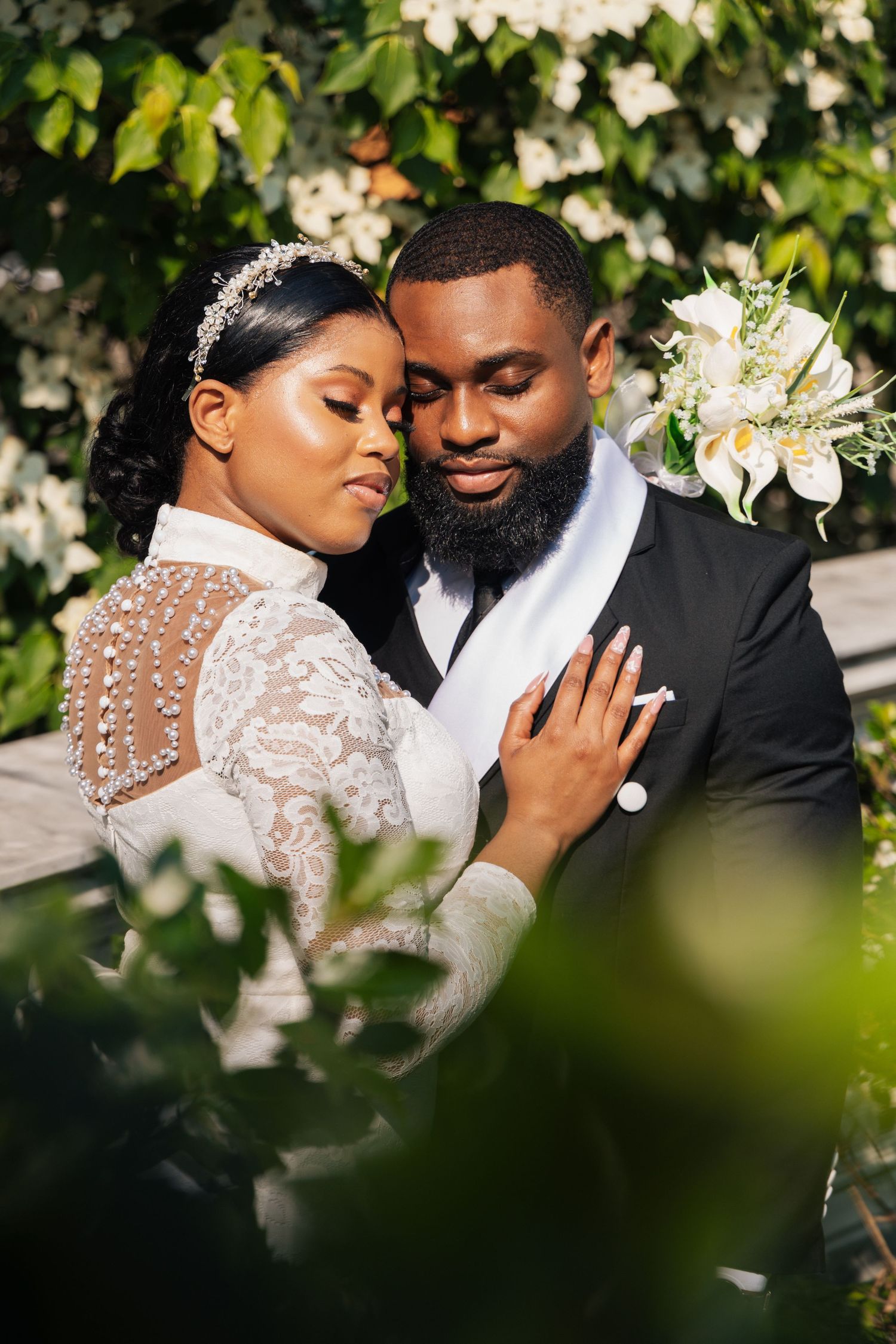 Black bride and groom embracing near white flowering bushes, bride in lace gown with pearl headpiece.