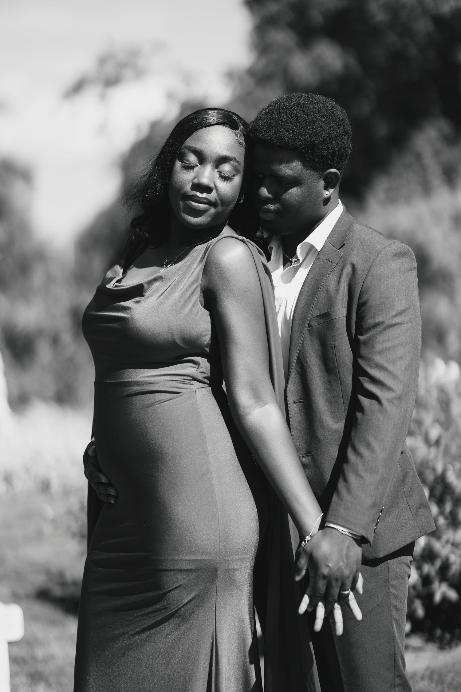 Black and white photo of a couple embracing outdoors, woman in elegant dress, man in suit holding hands tenderly.