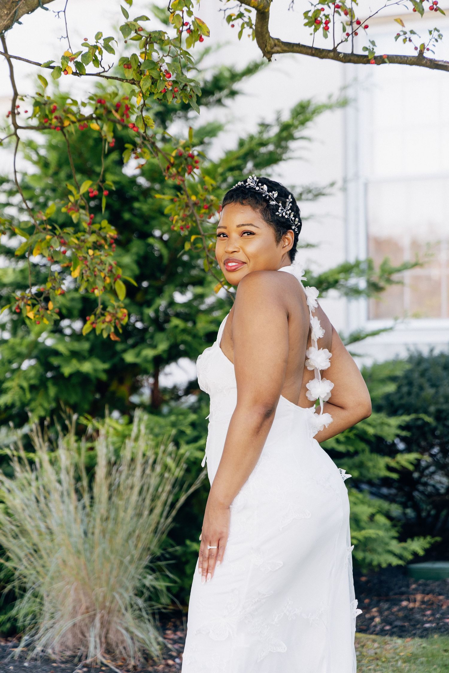 Bride in backless white lace gown with floral details poses outdoors near berry tree.