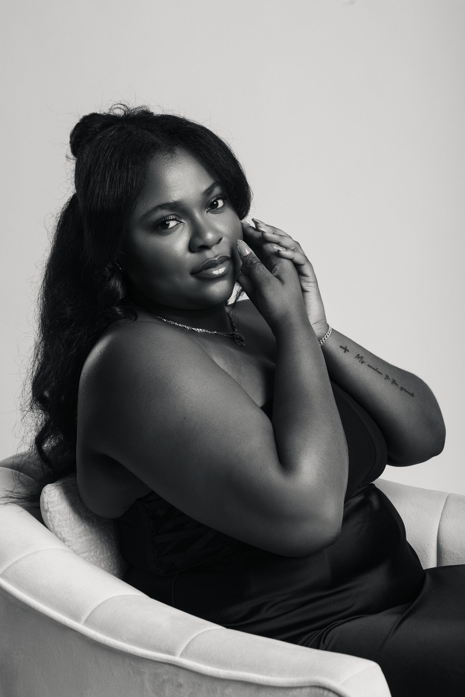 Black and white portrait of a woman sitting in a chair, posing gracefully with hand near face, wearing a sleeveless top.