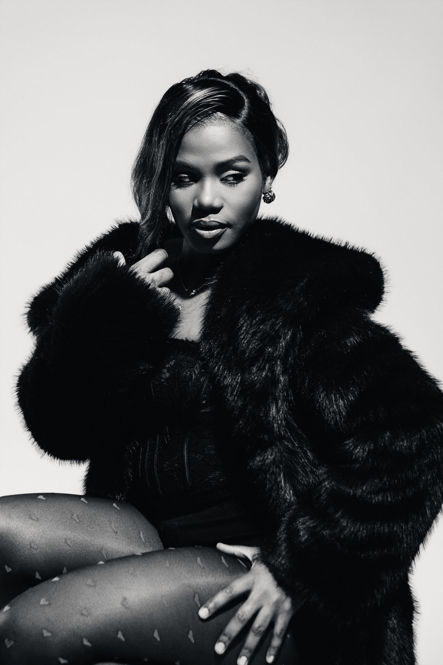 Black and white photo of a woman in a fur coat sitting confidently in patterned tights.