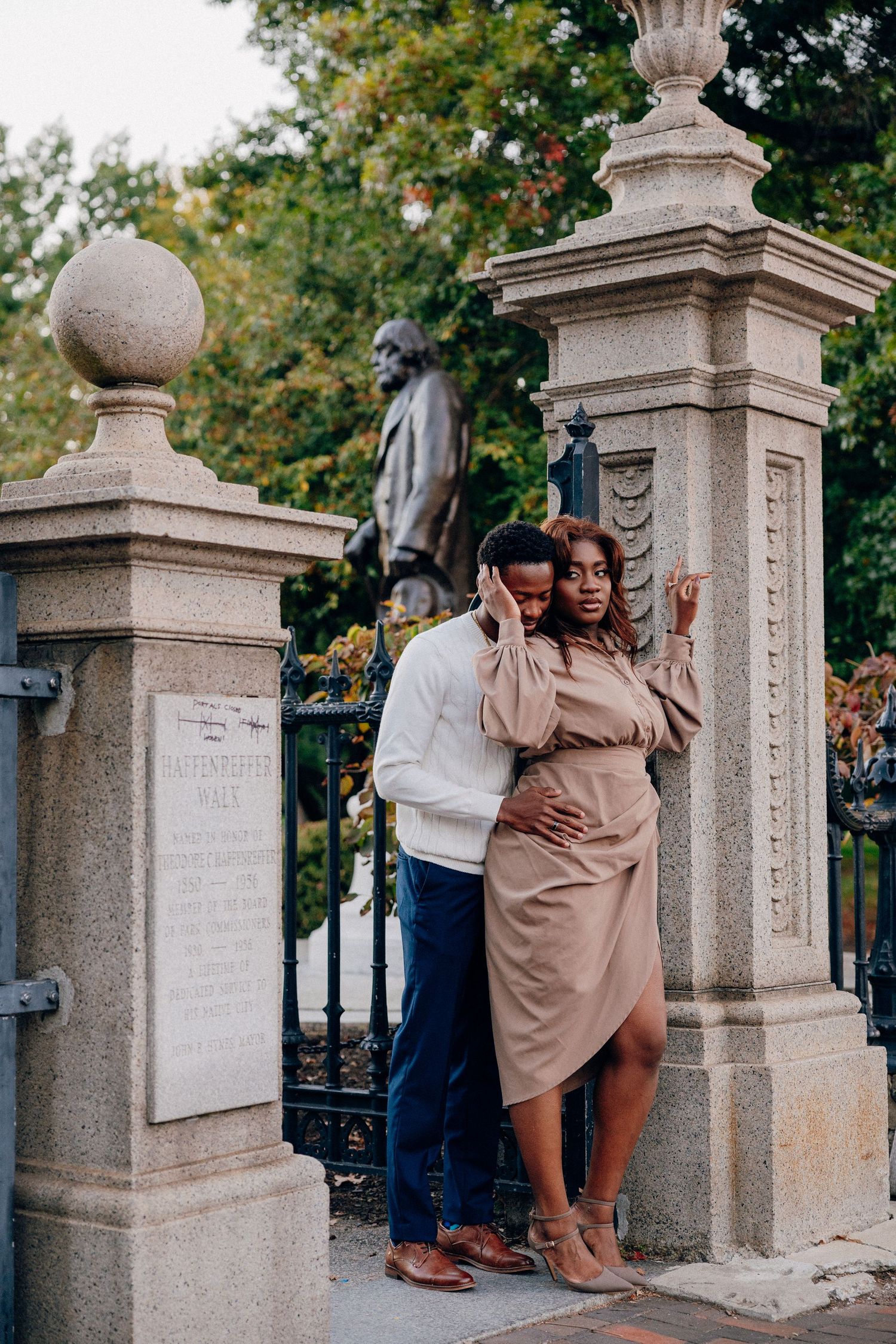 A couple embraces romantically by stone gate pillars with a bronze statue and lush green trees in the background.