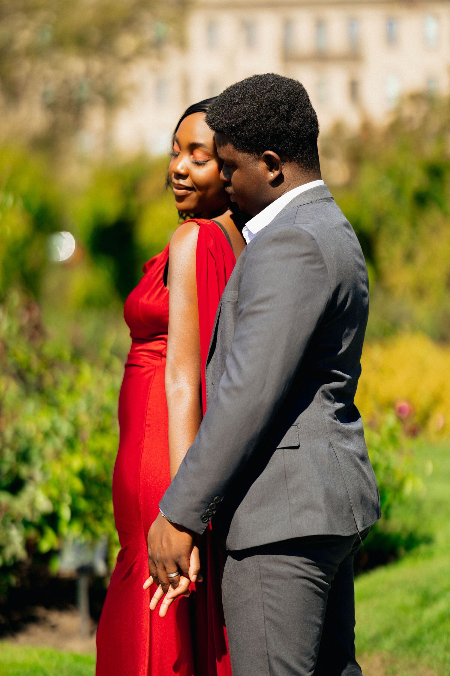 A couple in formal wear, woman in red dress and man in gray suit, holding hands in a lush green garden.