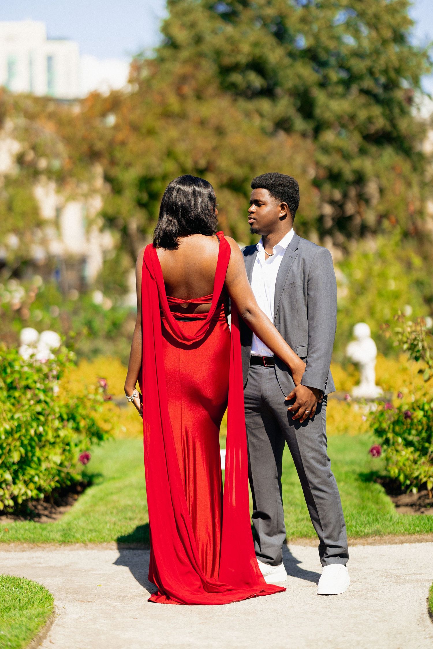 Elegant Black couple posing in garden, woman in red cape gown, man in gray suit, lush greenery background.