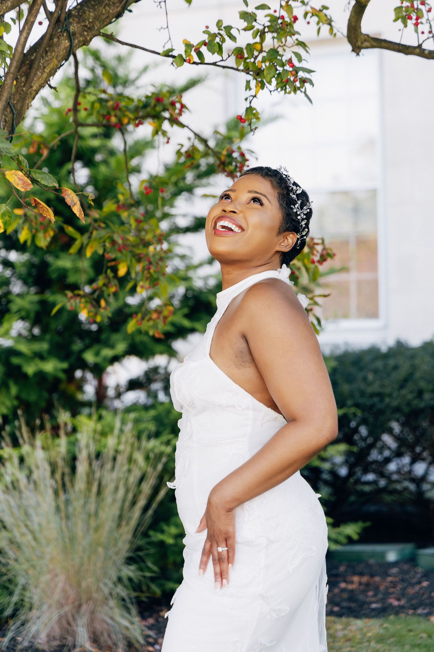 Smiling bride in white halter wedding dress standing beneath flowering tree in garden.