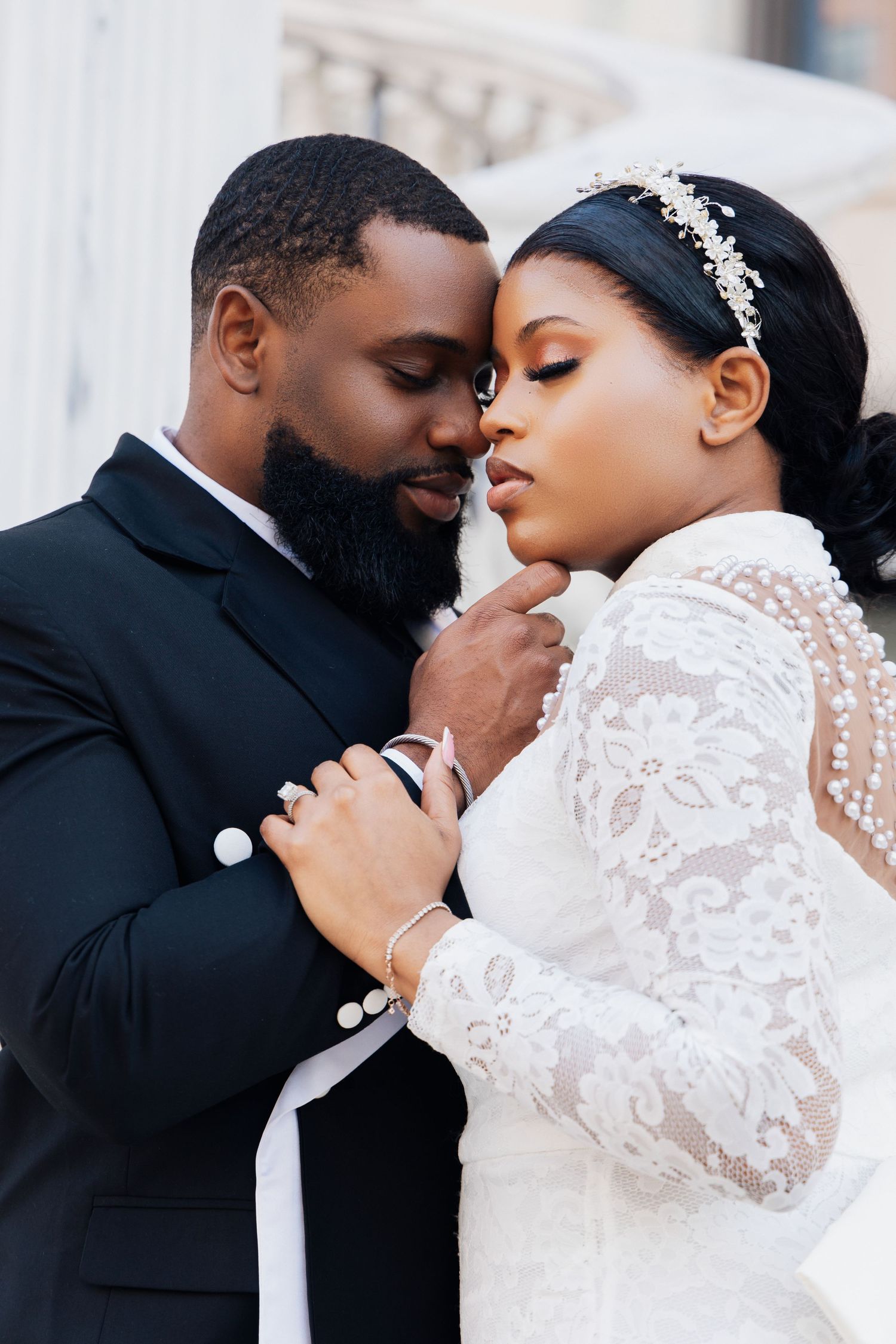 A Black bride and groom share an intimate moment, she in lace gown with pearl headband, he in black suit.