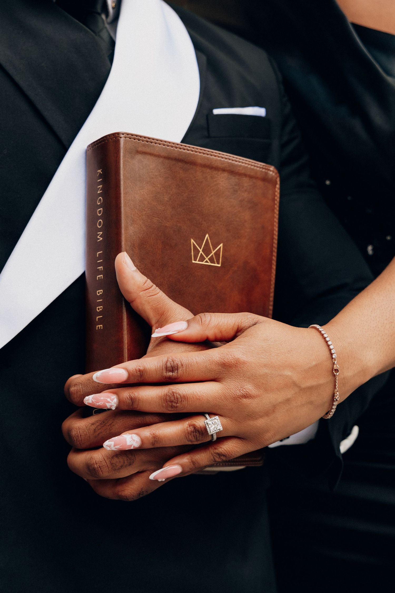 Hands holding a brown leather Kingdom Life Bible with a gold crown emblem, featuring a diamond ring and gold bracelet.
