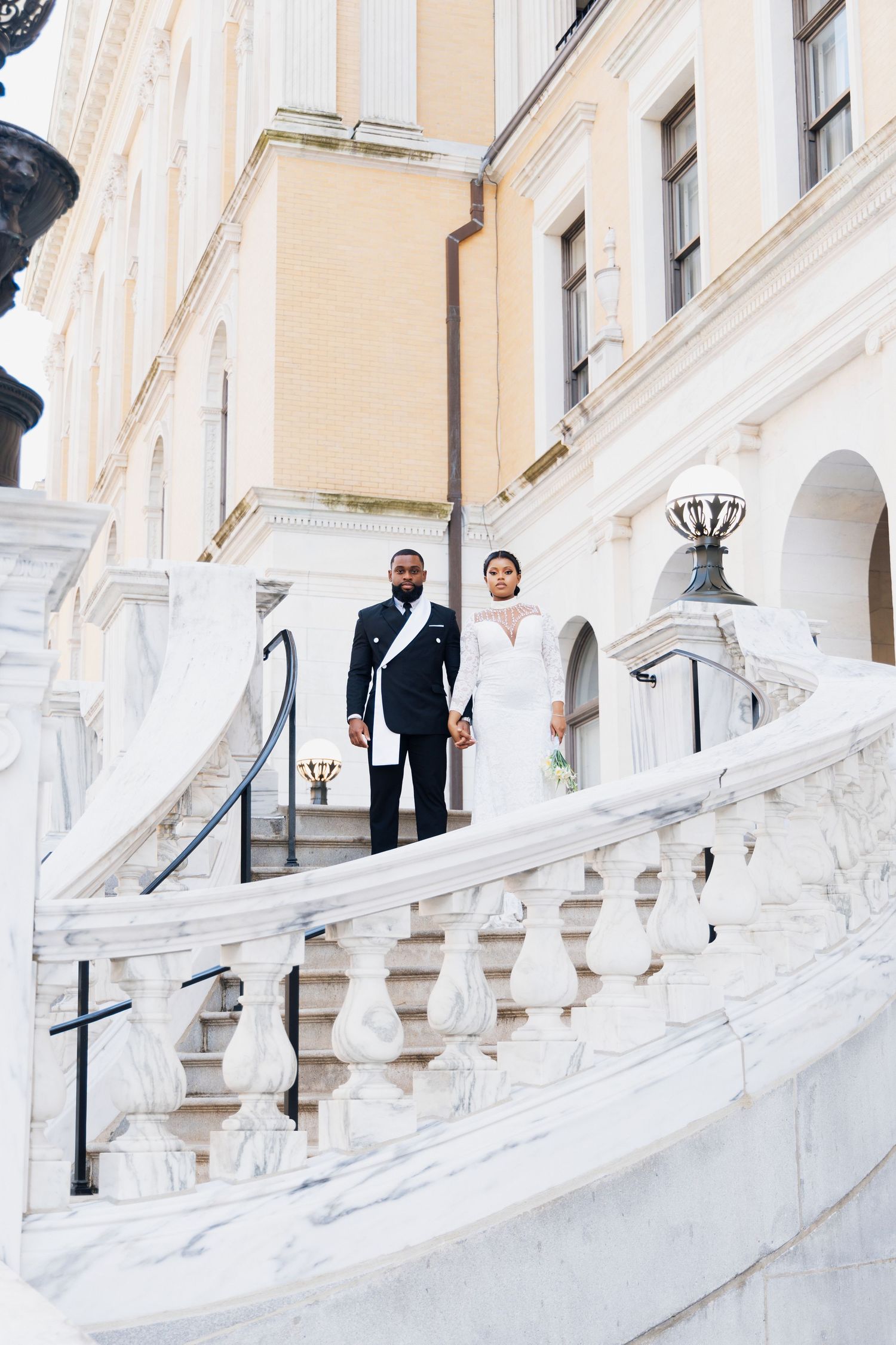 Elegant bride and groom in formal attire standing on grand marble staircase of a classical white building.