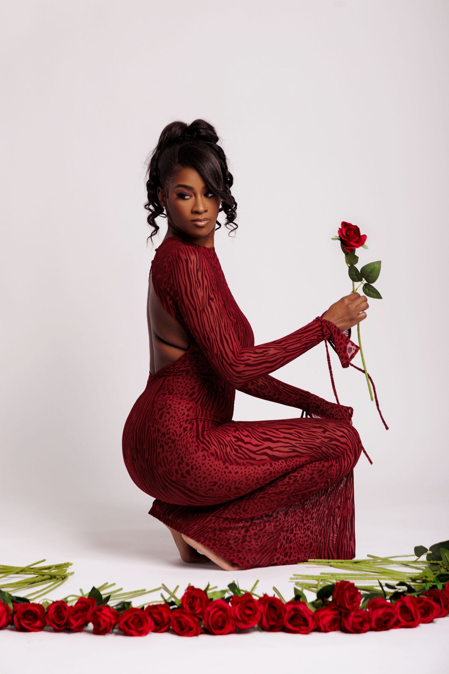 Woman in red backless gown holding a single red rose, surrounded by roses on white background.