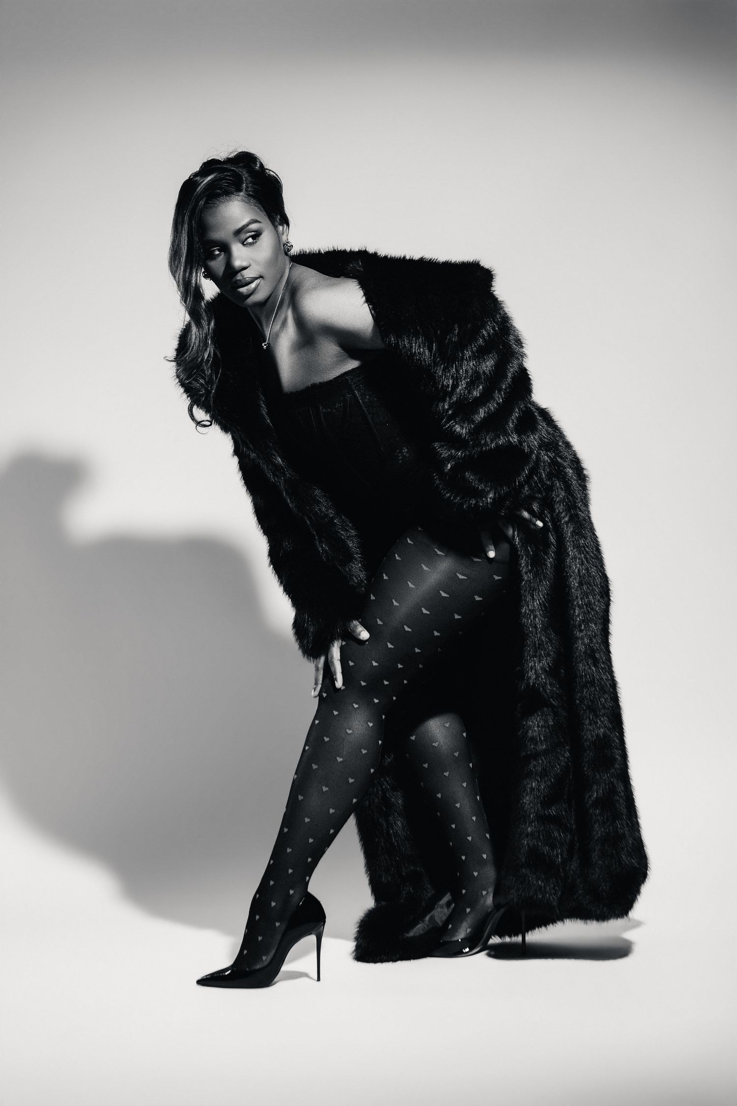 Black and white photo of a woman in a fur coat, patterned tights, and heels posing confidently in a studio.