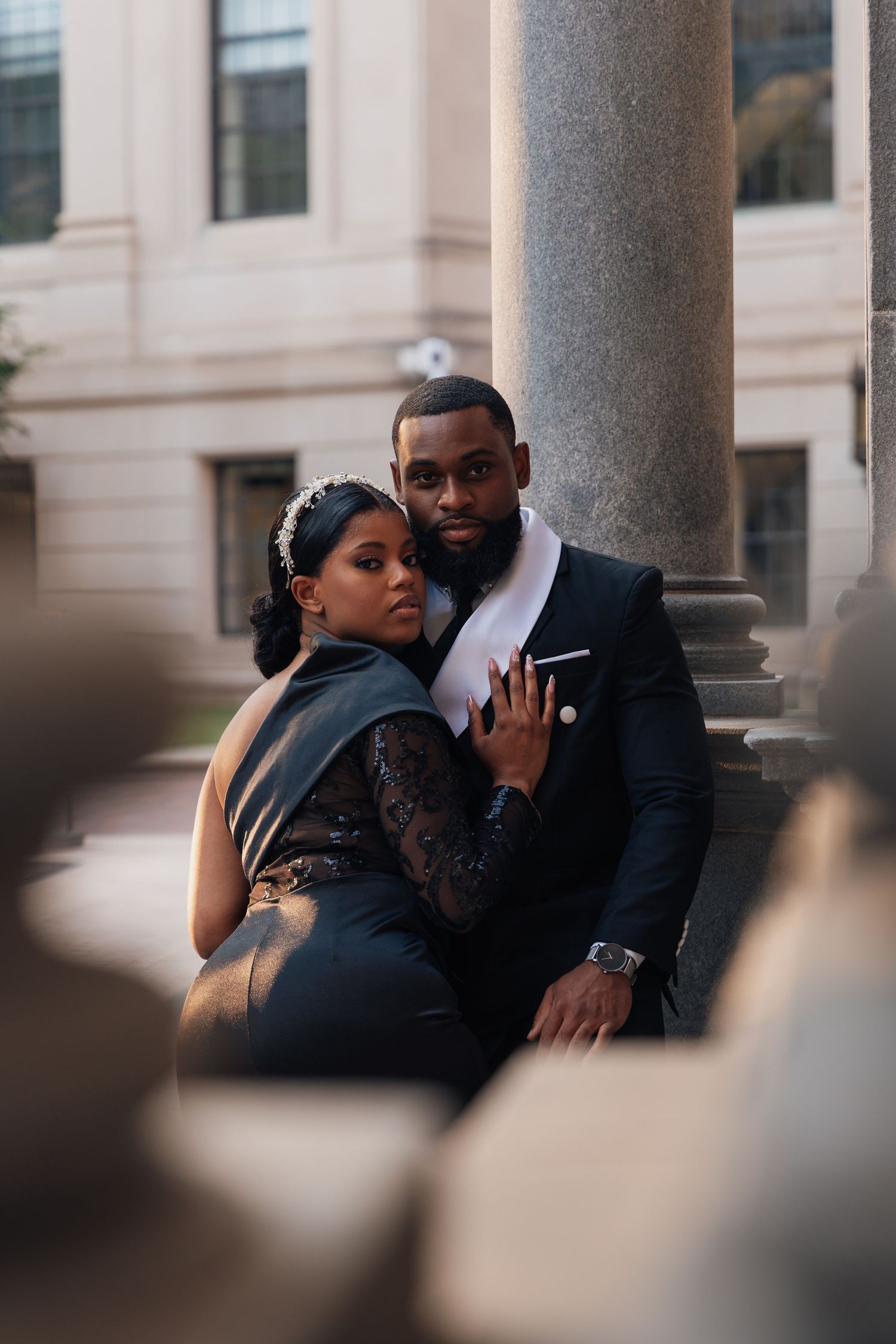 Elegant Black couple in formal attire posing by stone columns, woman in lace dress embracing suited man.