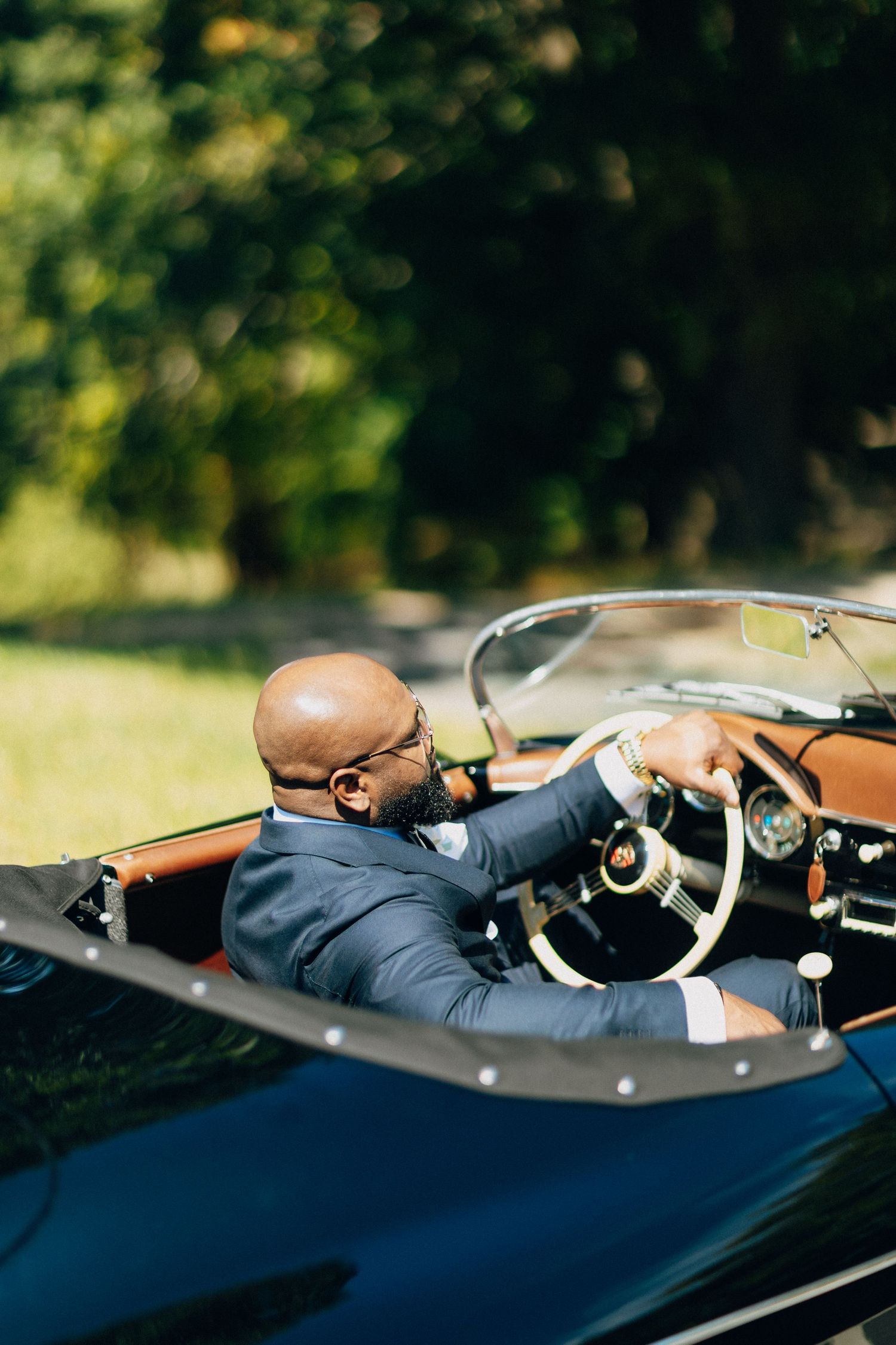 Groom in a navy suit driving a classic vintage convertible car on a sunny day outdoors.