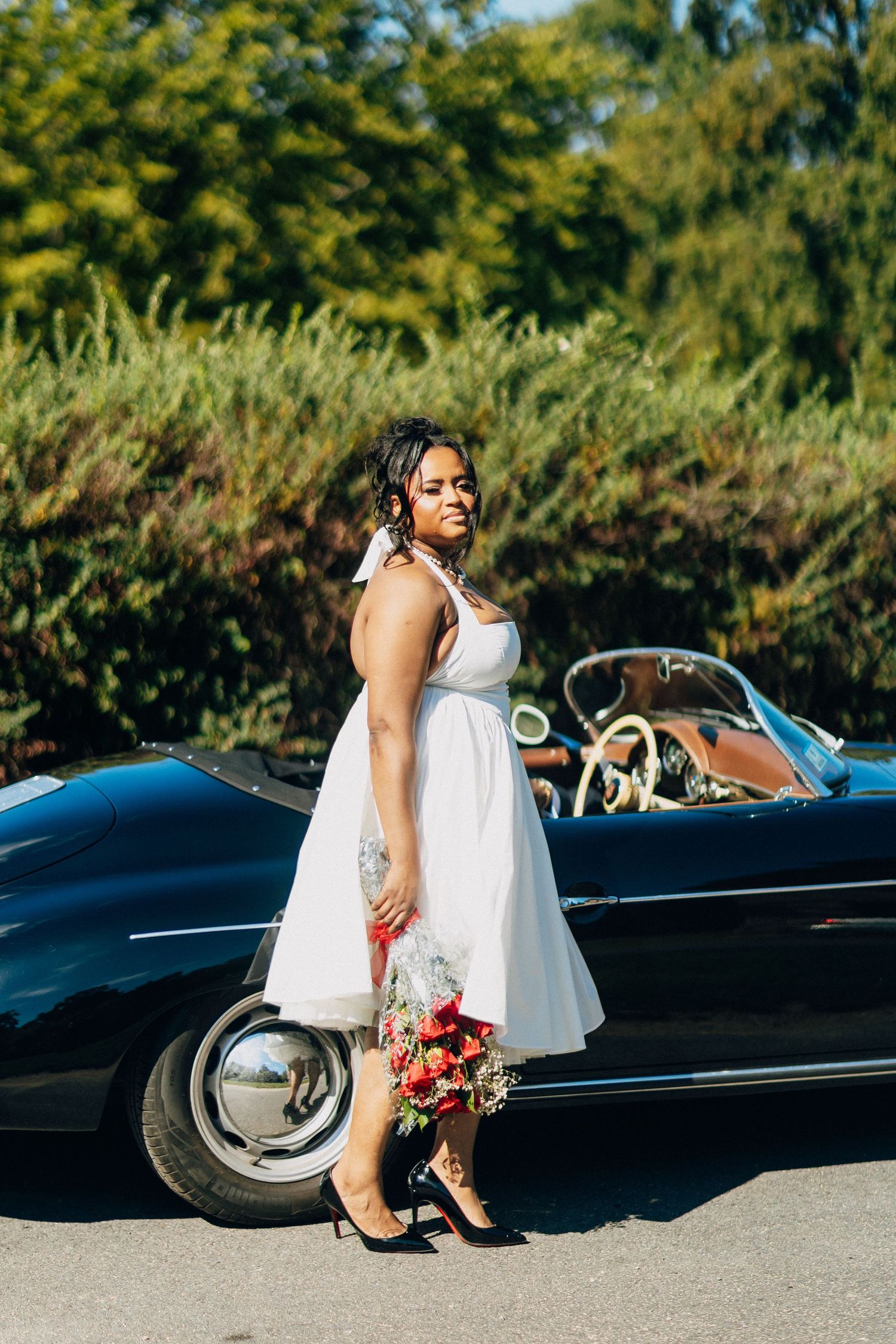 Bride in white tea-length dress poses with bridal bouquet beside a vintage black convertible car.