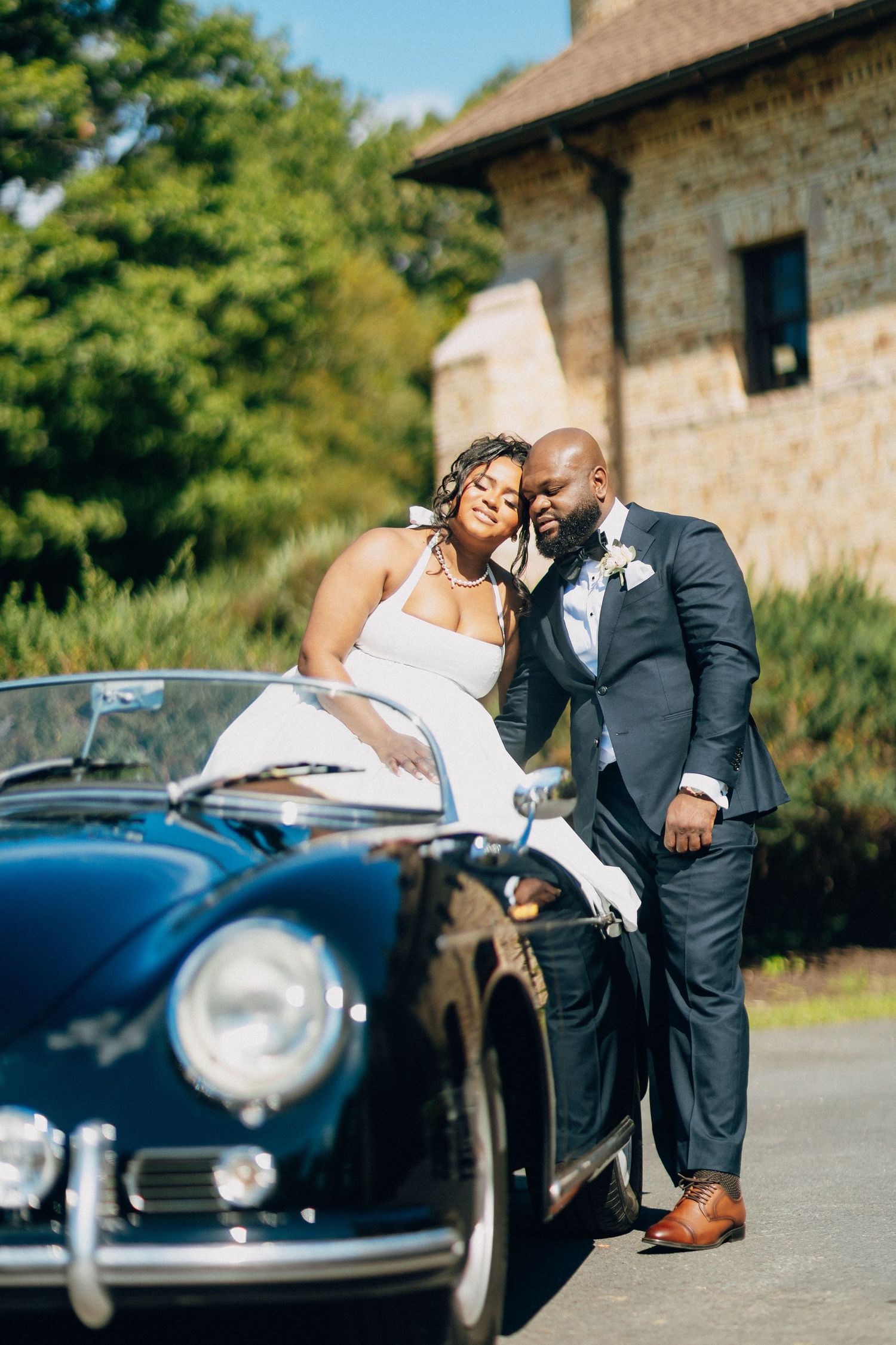 Bride and groom pose elegantly beside a classic vintage Porsche convertible on their wedding day outdoors.