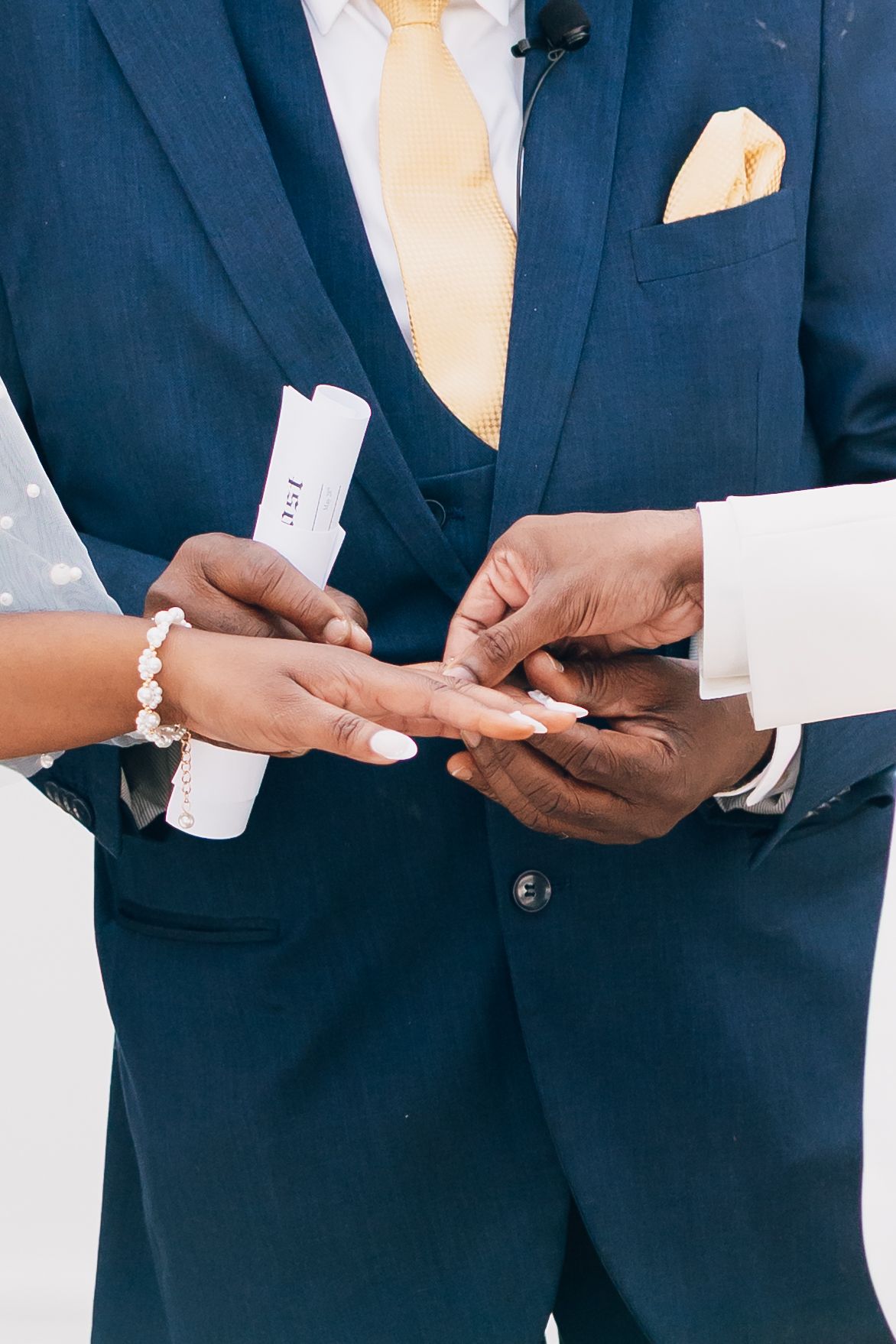 Groom in navy suit placing wedding ring on bride's finger during ceremony, bride holding vows scroll.