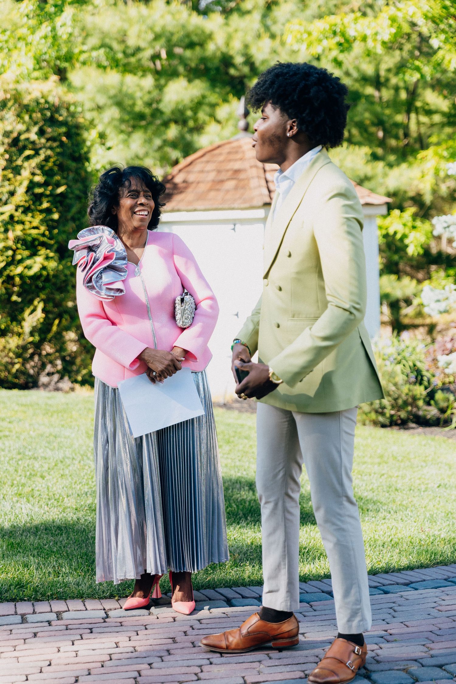 Elegant woman in pink jacket and man in cream suit conversing outdoors at a garden event.
