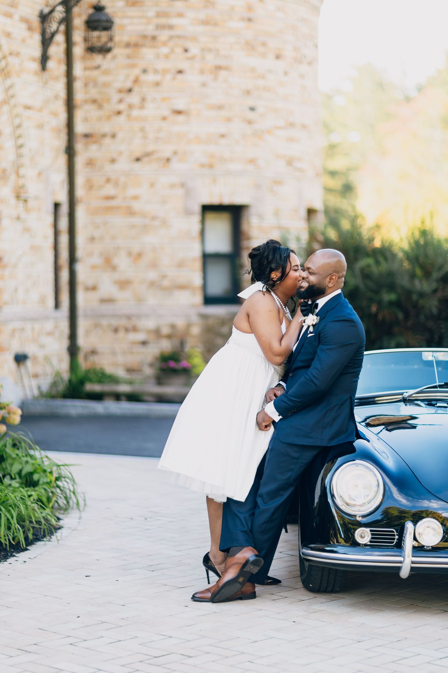 Newlywed couple shares a romantic kiss leaning against a vintage black Porsche outside a stone building.
