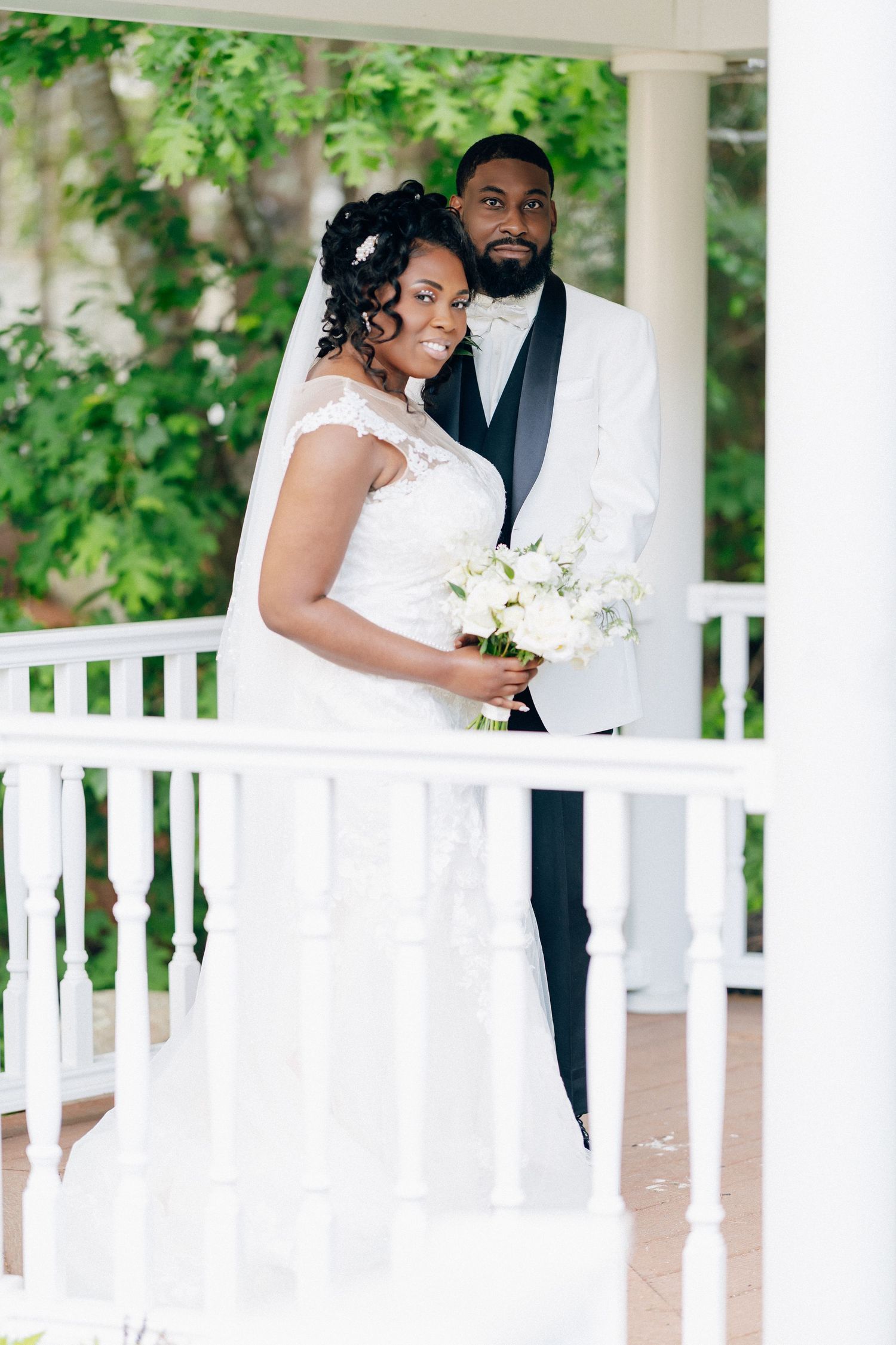 Bride and groom posing on white gazebo, bride in lace gown holding white bouquet, groom in white tuxedo jacket.