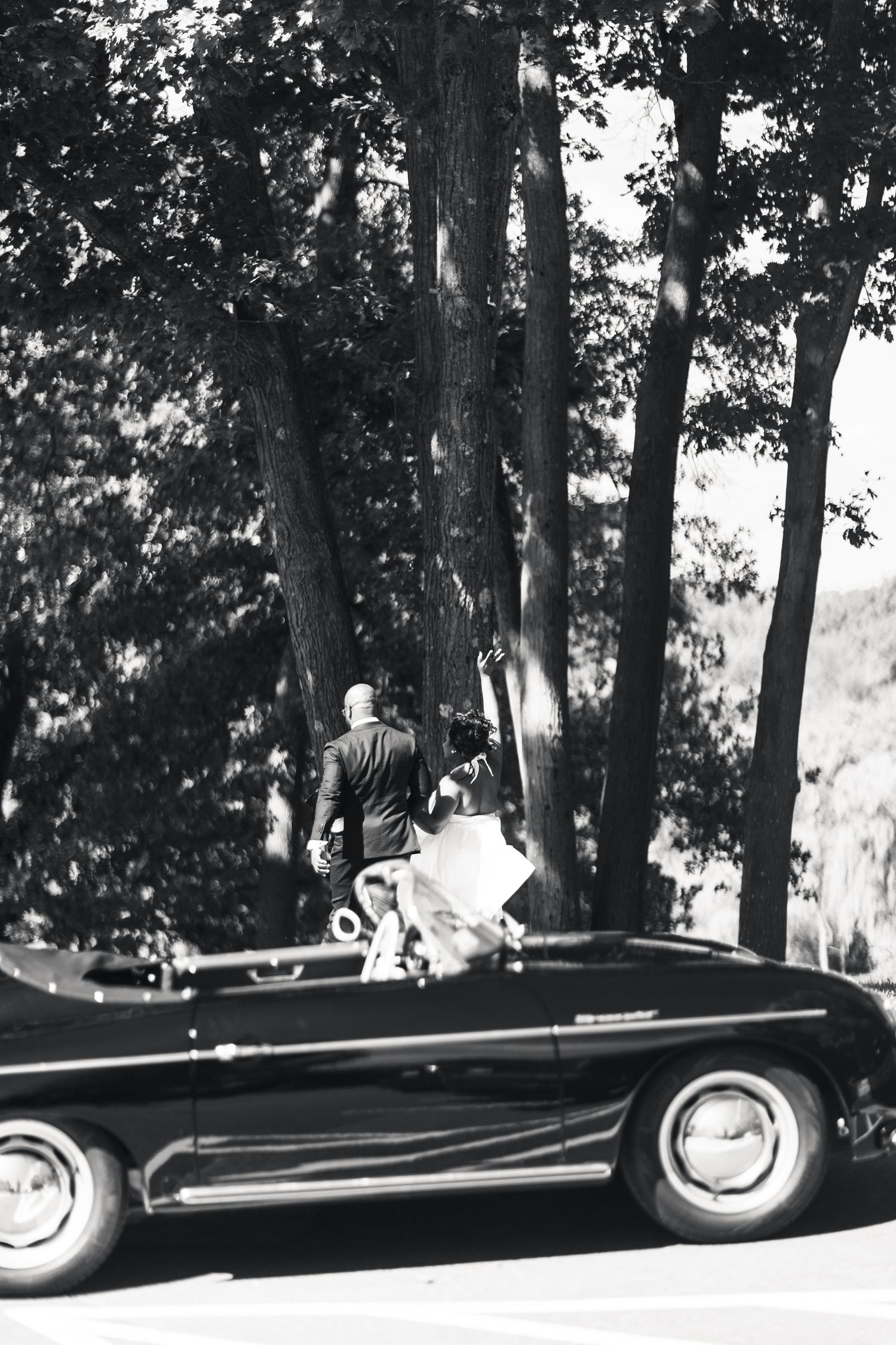 Black and white photo of bride and groom walking beside a vintage convertible among trees.