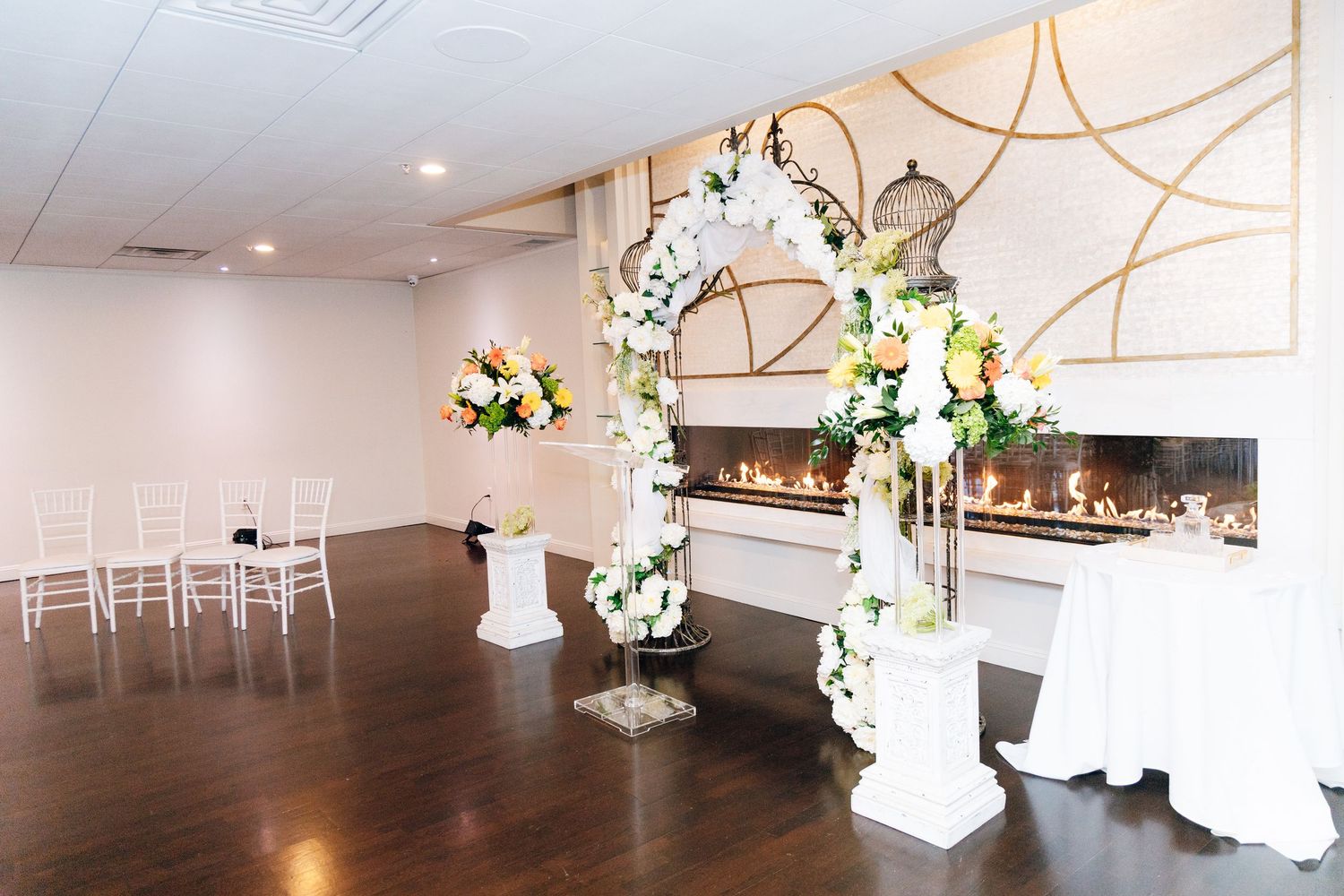 Elegant wedding ceremony venue with floral arch, white chairs, and a modern fireplace adorned with gold geometric decor.