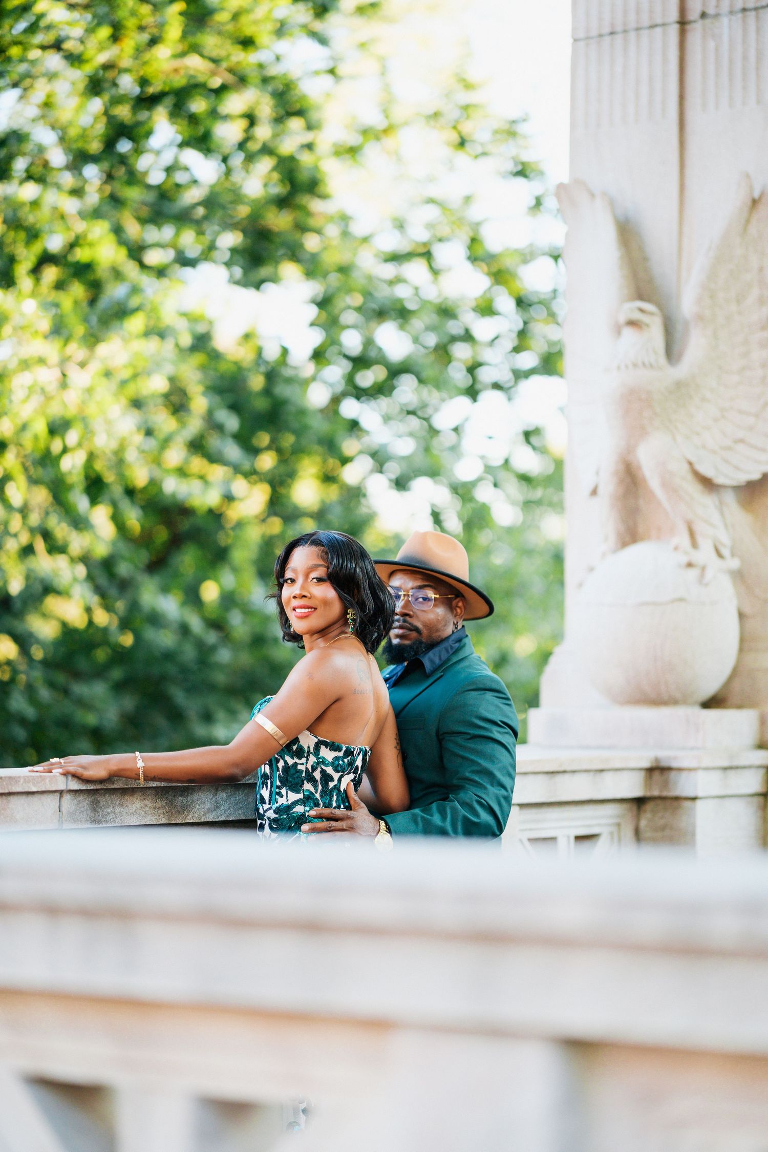 Elegant Black couple posing near stone monument with eagle sculpture in lush green park.