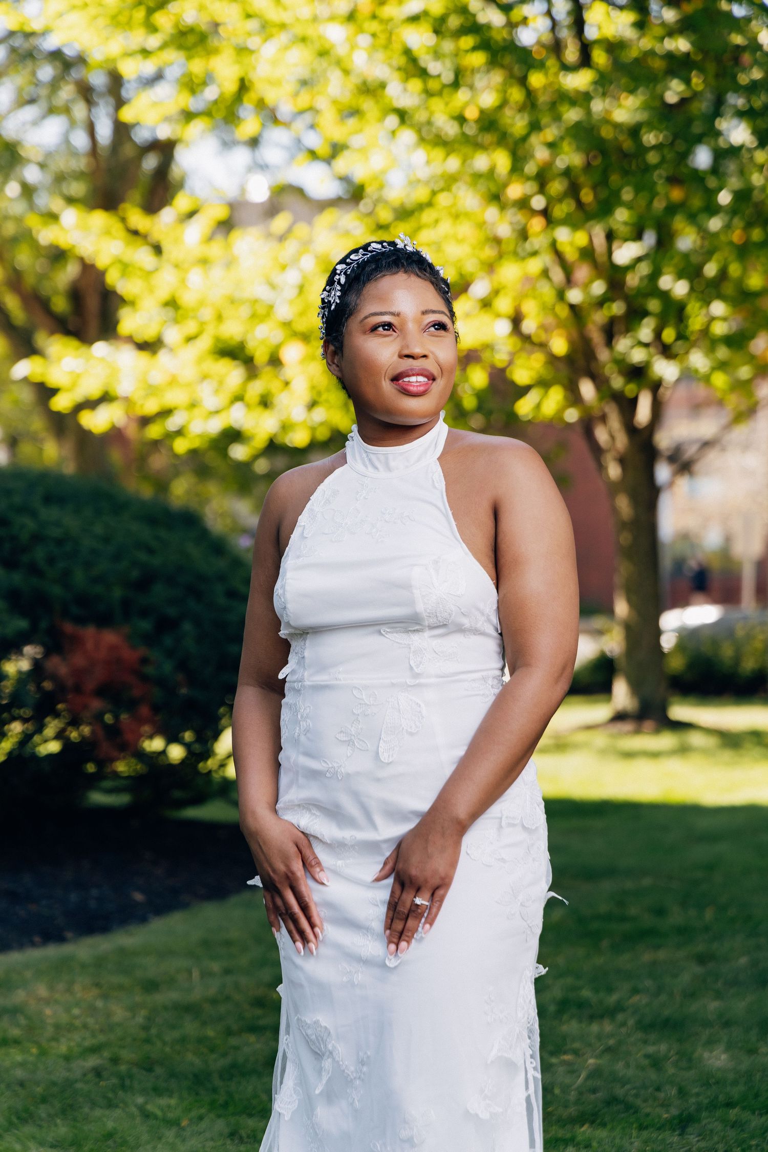 Radiant bride in white halter wedding dress with pearl headband poses in sunlit garden with lush green trees.
