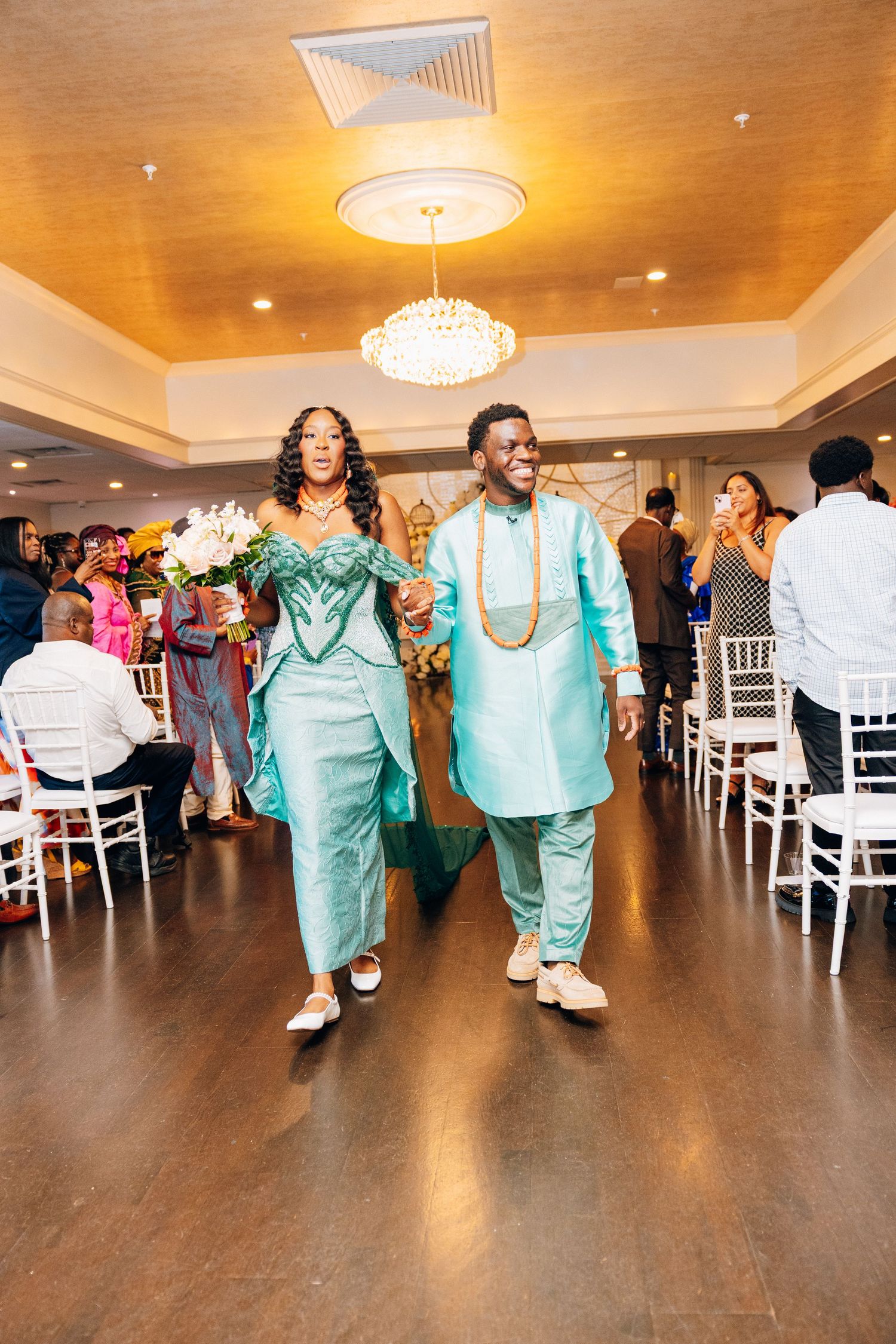 Couple in matching teal traditional African attire walks down the aisle at an elegant wedding reception venue.