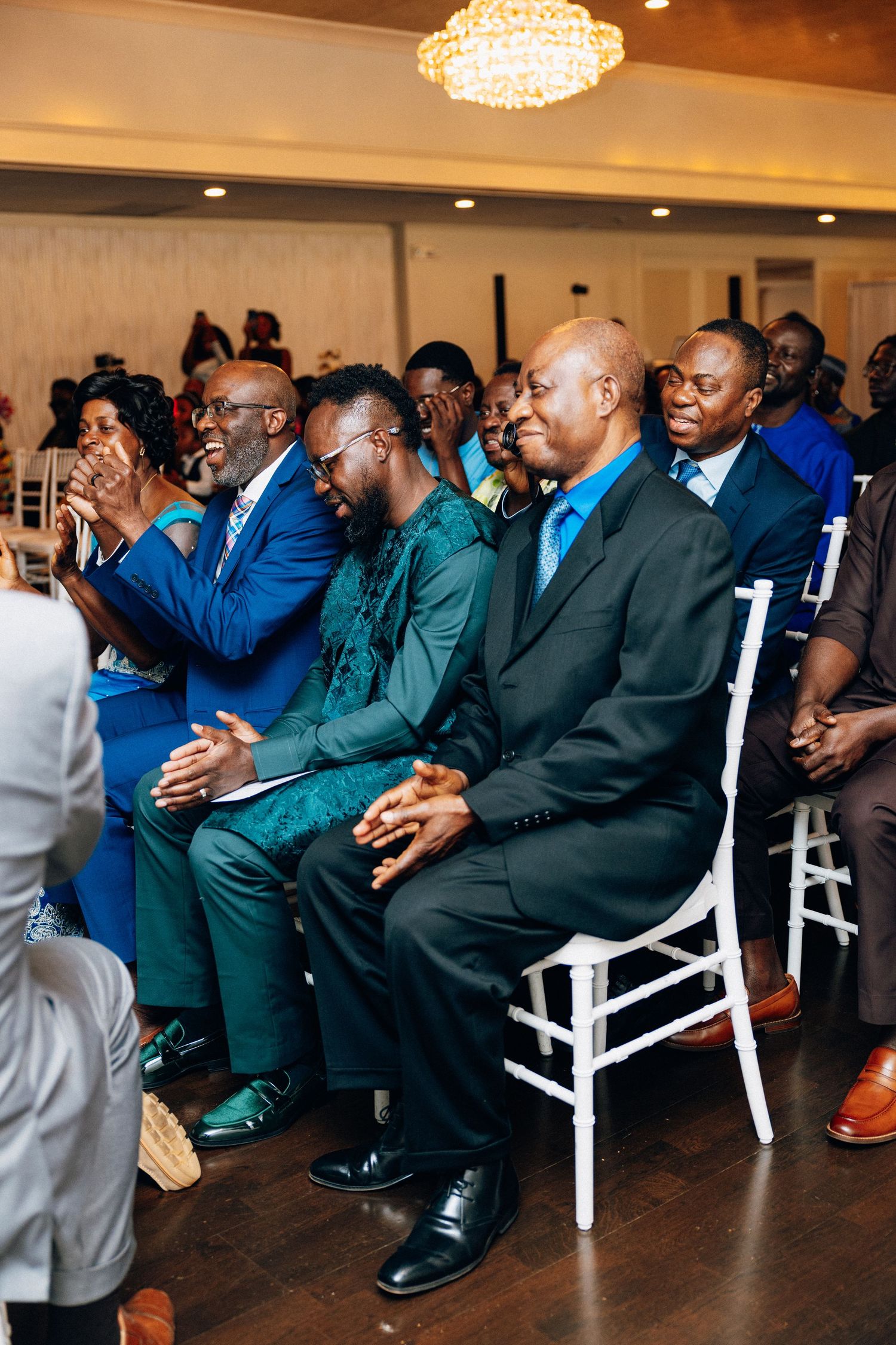 Wedding guests in colorful attire seated and smiling during ceremony in elegant venue.