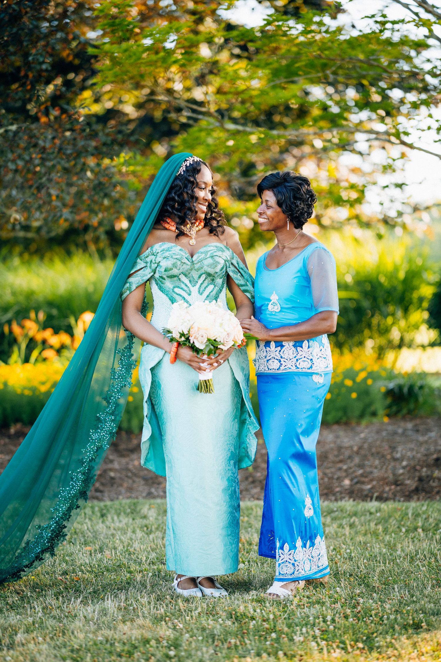 Bride in teal gown with veil holds white bouquet beside woman in blue dress, standing in lush garden.