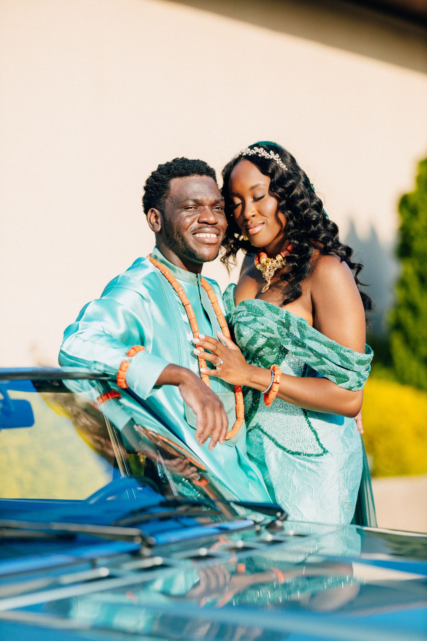 Nigerian couple in matching teal traditional wedding attire pose smiling beside vintage blue car.