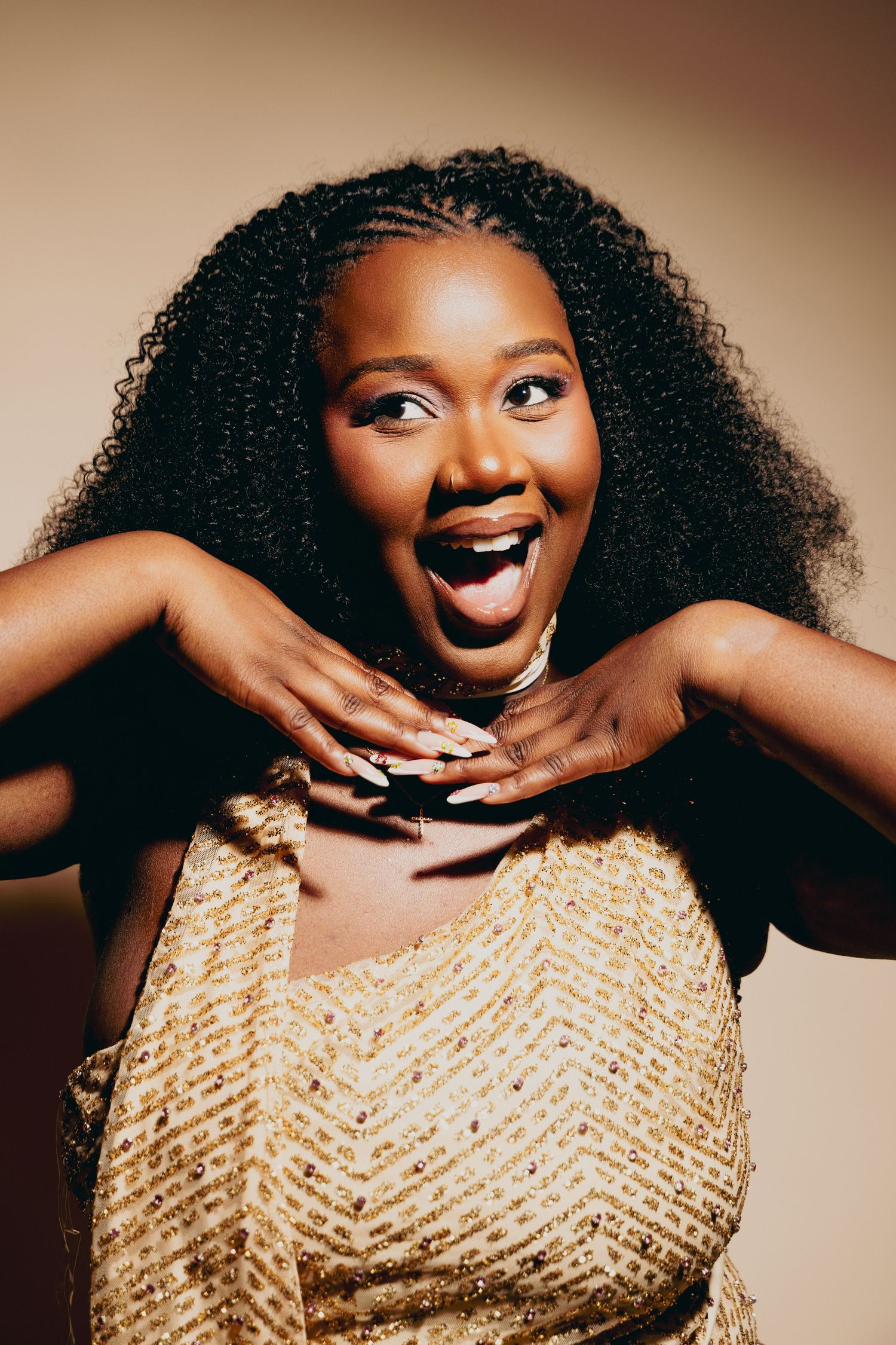 A joyful Black woman in a gold sequin dress with voluminous curly hair poses with hands near chin, mouth open in surprise.