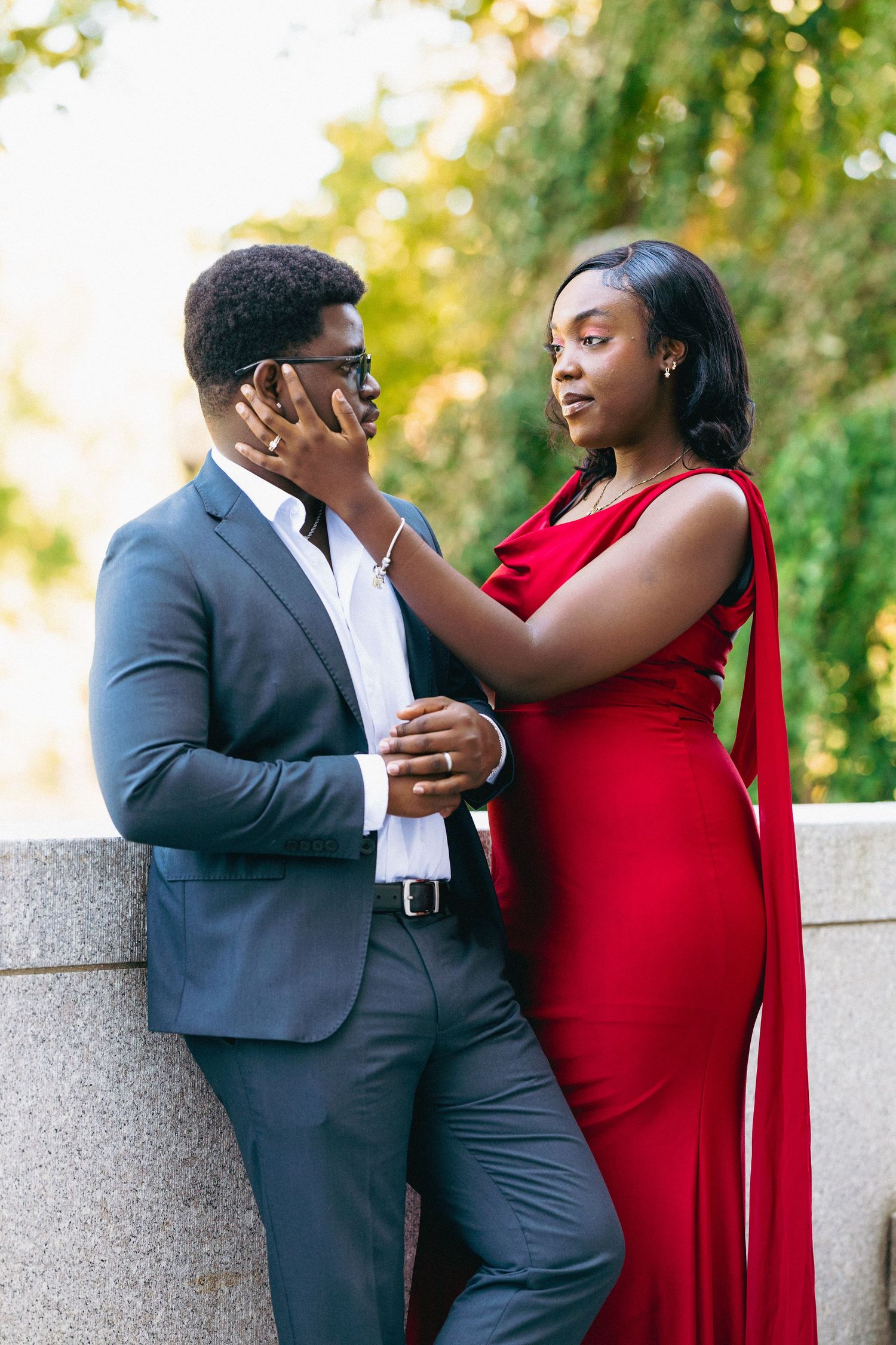 Elegant Black couple posing outdoors, man in gray suit with sunglasses, woman in red gown touching his face.