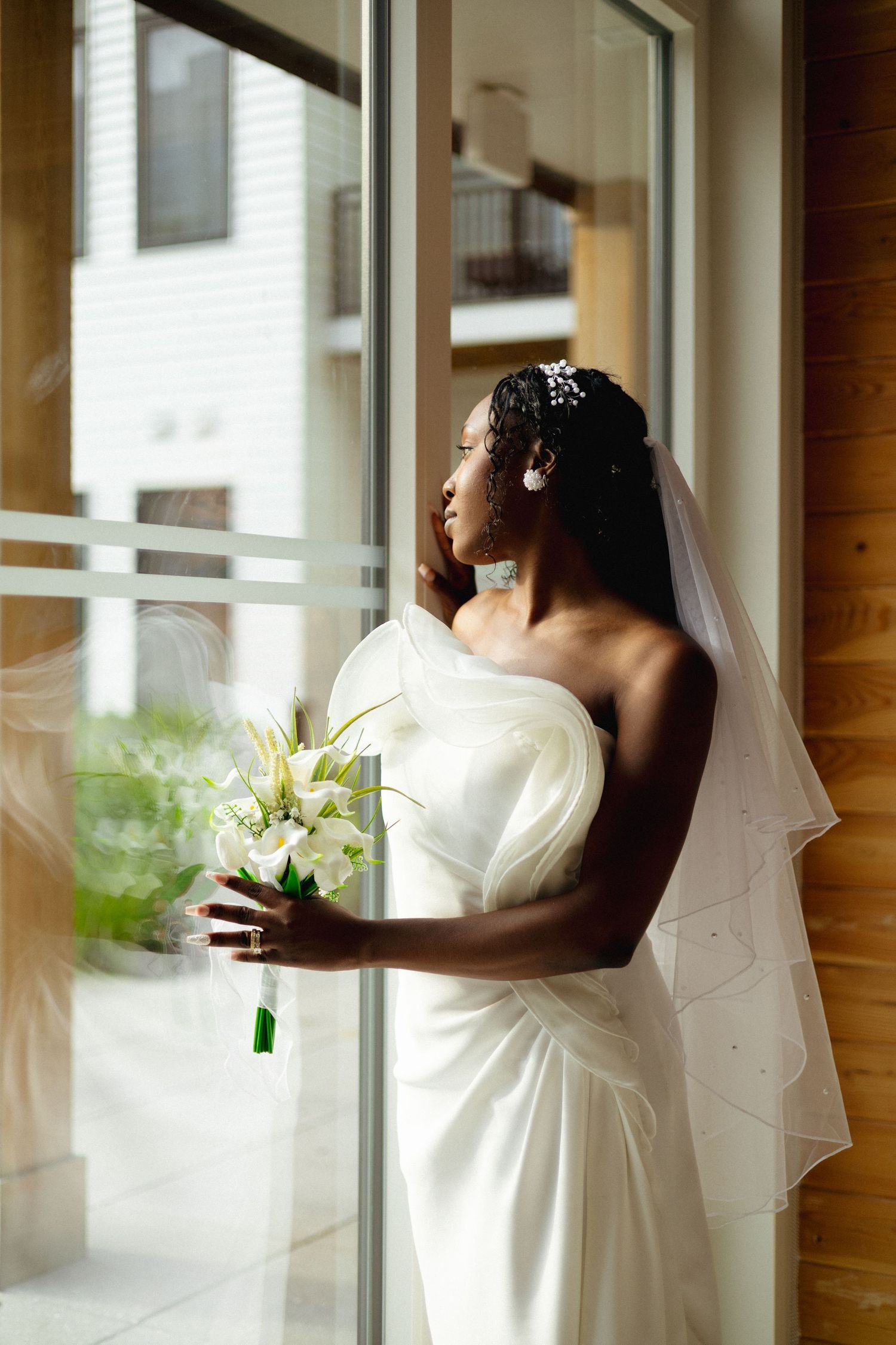 Elegant Black bride in white strapless gown and veil holds white floral bouquet while gazing out a sunlit window.
