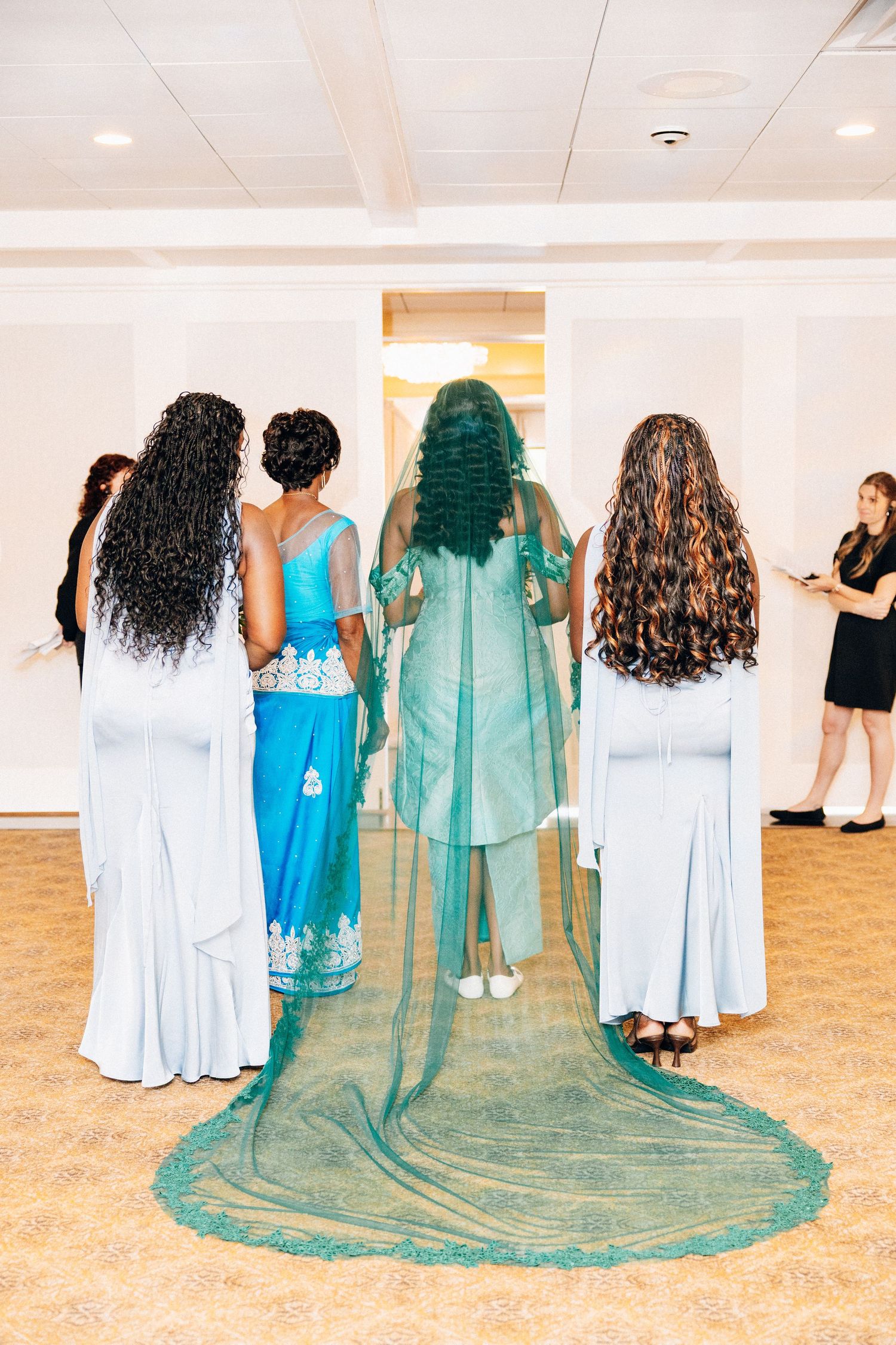 Bride in teal African gown with long matching veil stands with bridesmaids in light blue dresses viewed from behind.