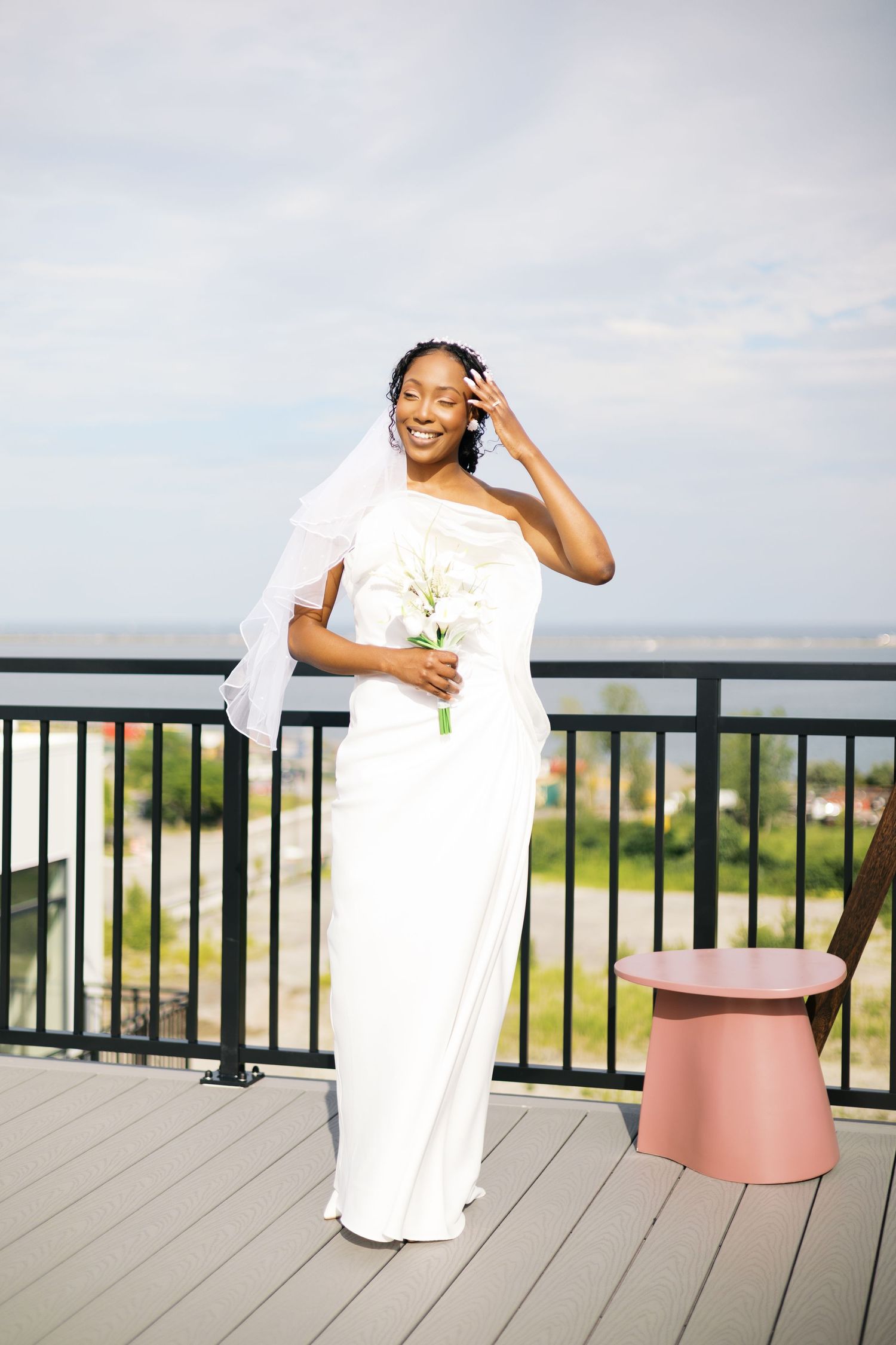 Smiling Black bride in white gown and veil holds bouquet on outdoor deck with scenic view.