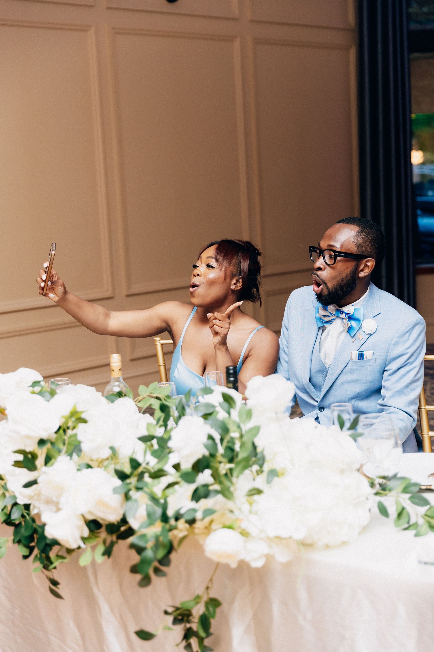 Bride and groom take a selfie at their wedding reception sweetheart table adorned with white floral arrangements.
