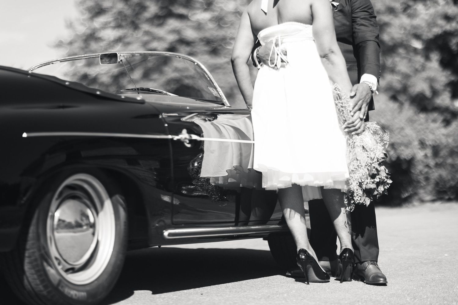 Black and white photo of bride and groom standing beside a classic vintage convertible wedding car.