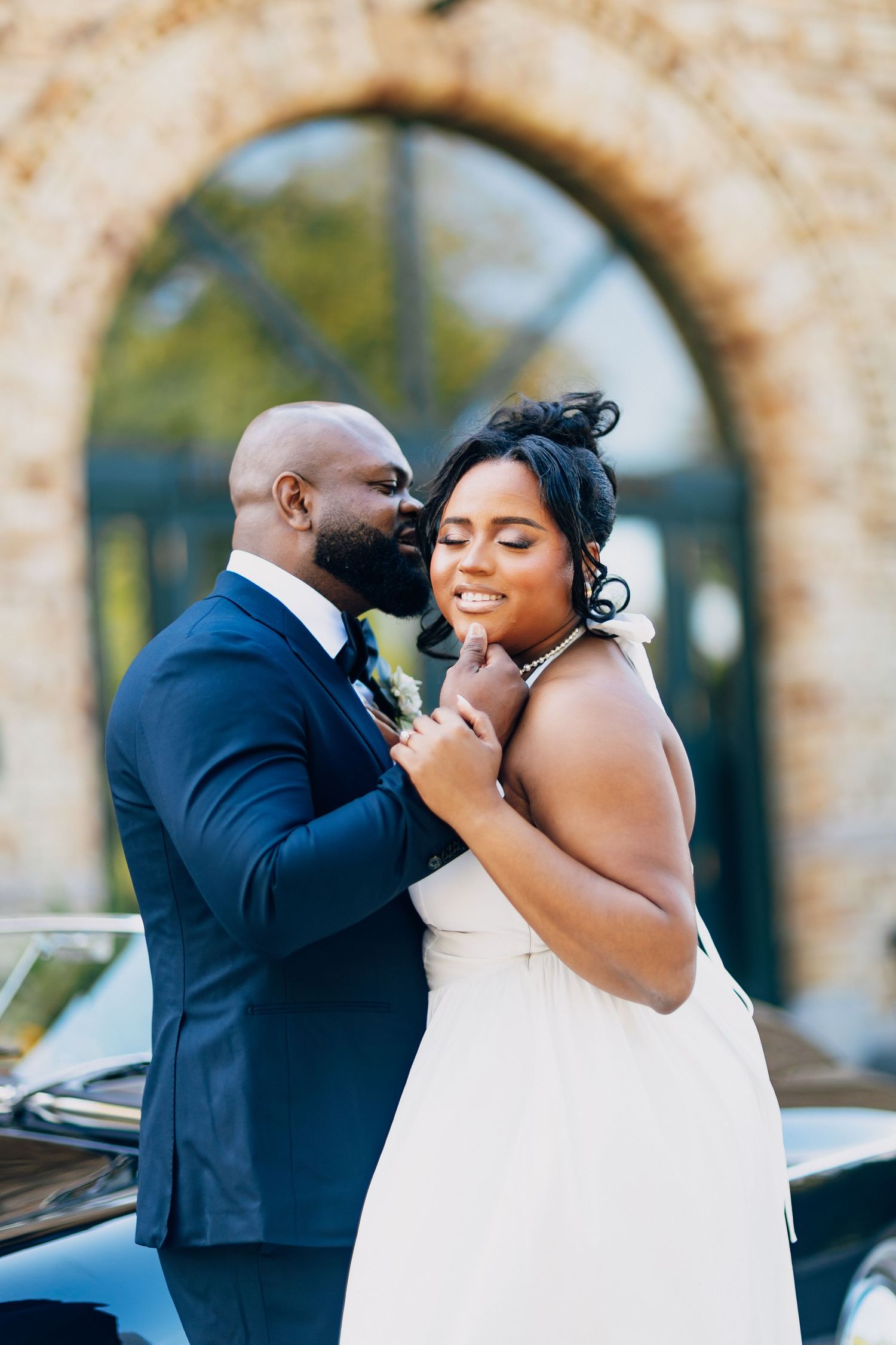 Black groom in navy suit whispering to smiling bride in white dress beside a vintage car outdoors.