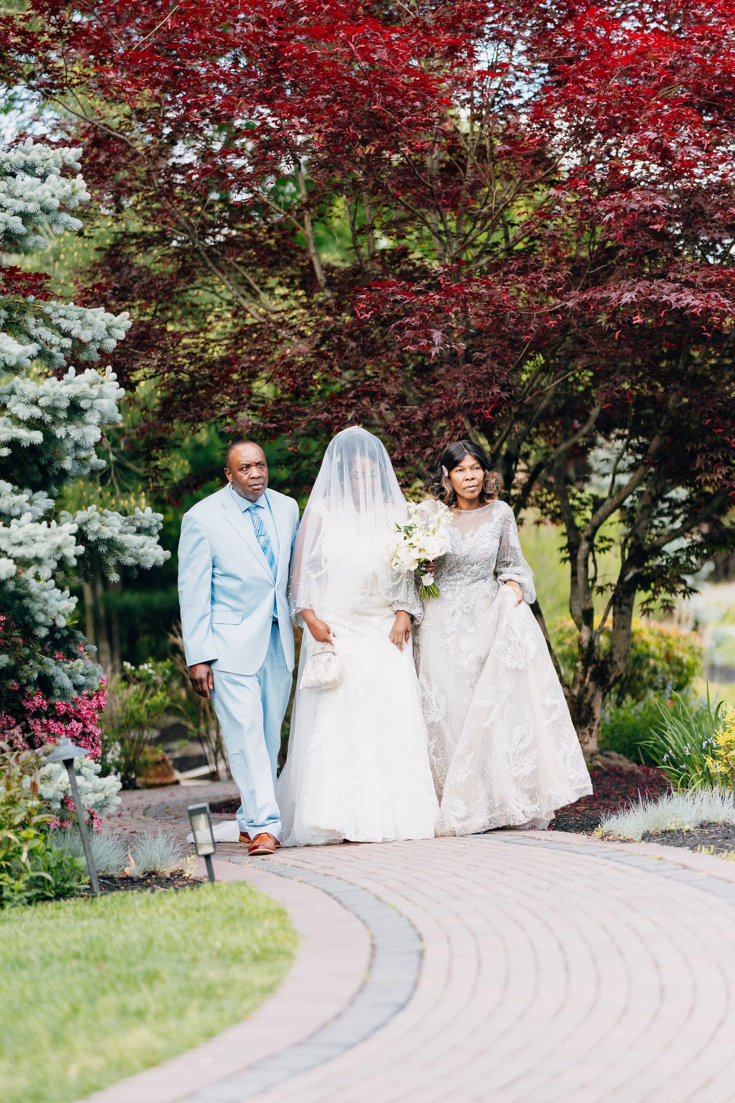 Bride walks down garden path flanked by parents in light blue suit and silver gown amid red maple trees.