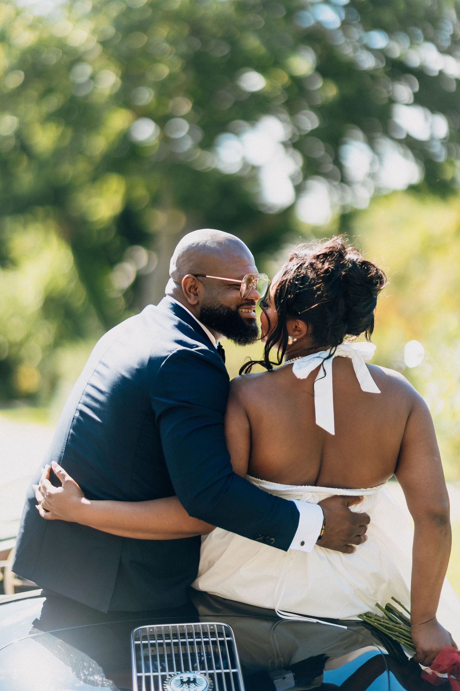 Groom in navy suit kissing bride on cheek while sitting together on a vintage convertible car.
