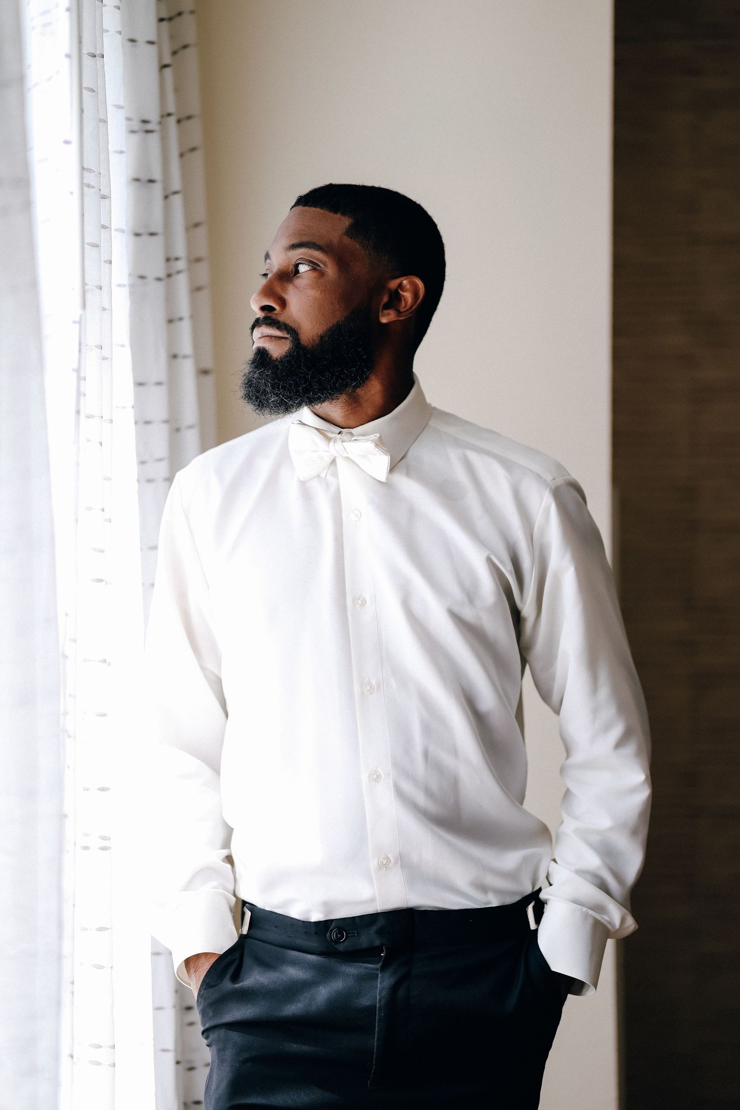 Handsome bearded Black groom in white dress shirt and bow tie standing by window.