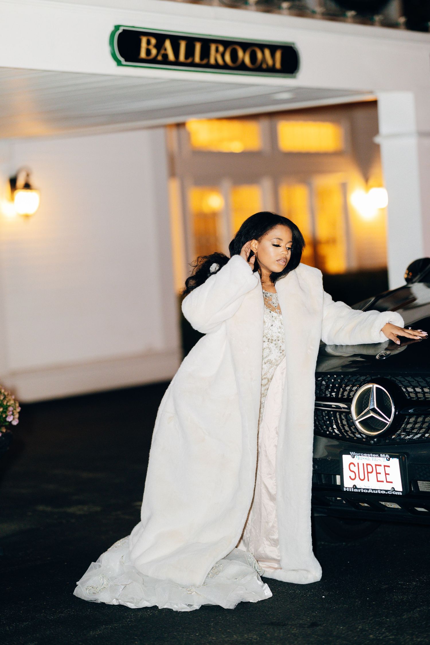 Elegant woman in white fur coat and gown posing beside a Mercedes outside a ballroom at night.