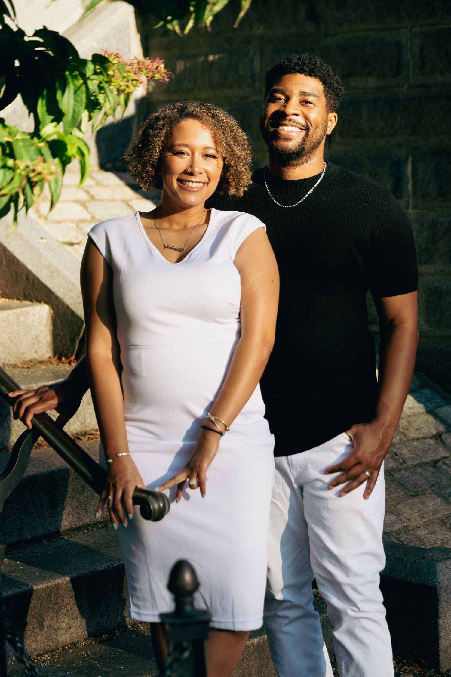 A smiling couple poses on stone steps, woman in white dress and man in black shirt with white pants.