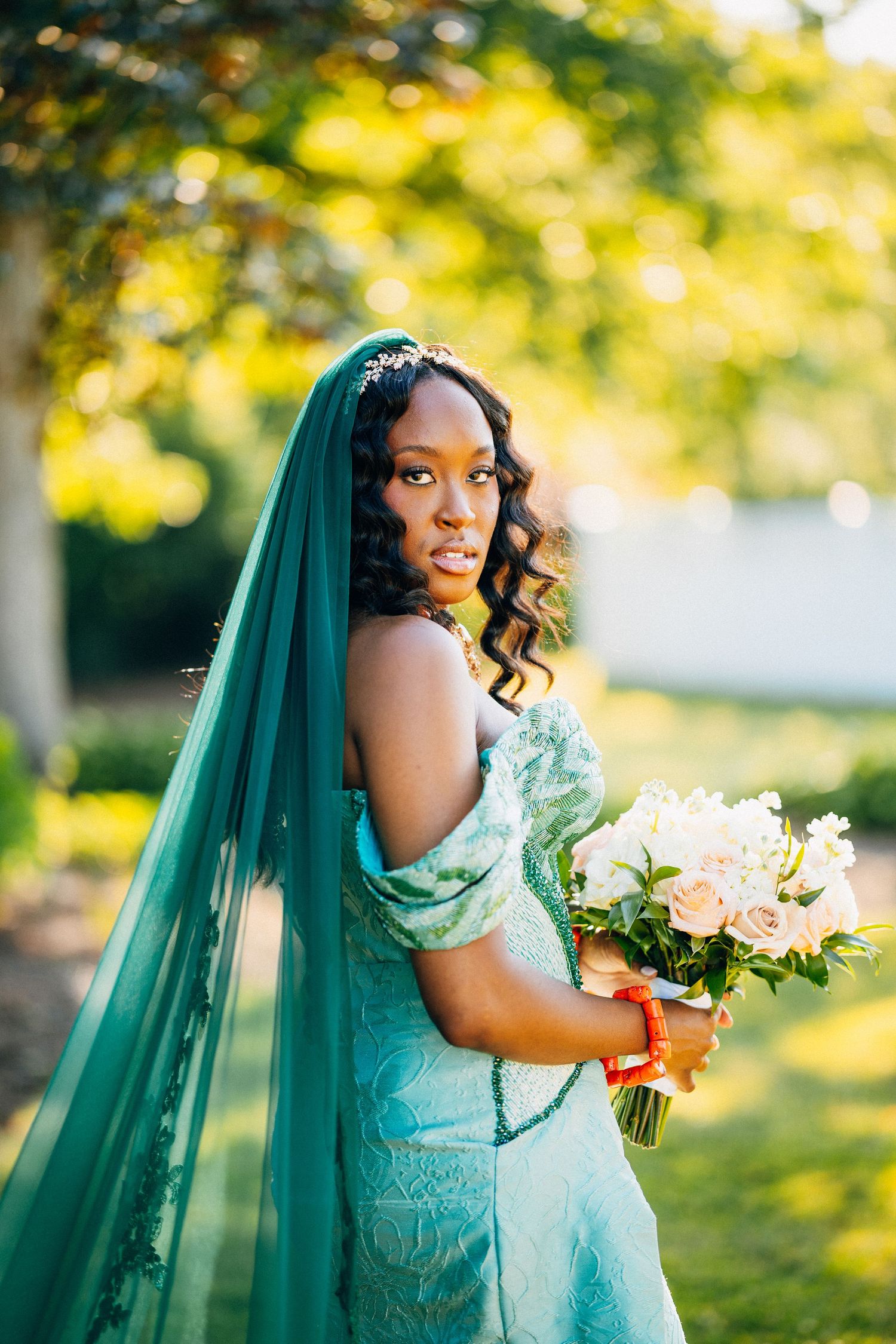 Black bride in teal lace gown and matching veil holds blush bouquet outdoors.